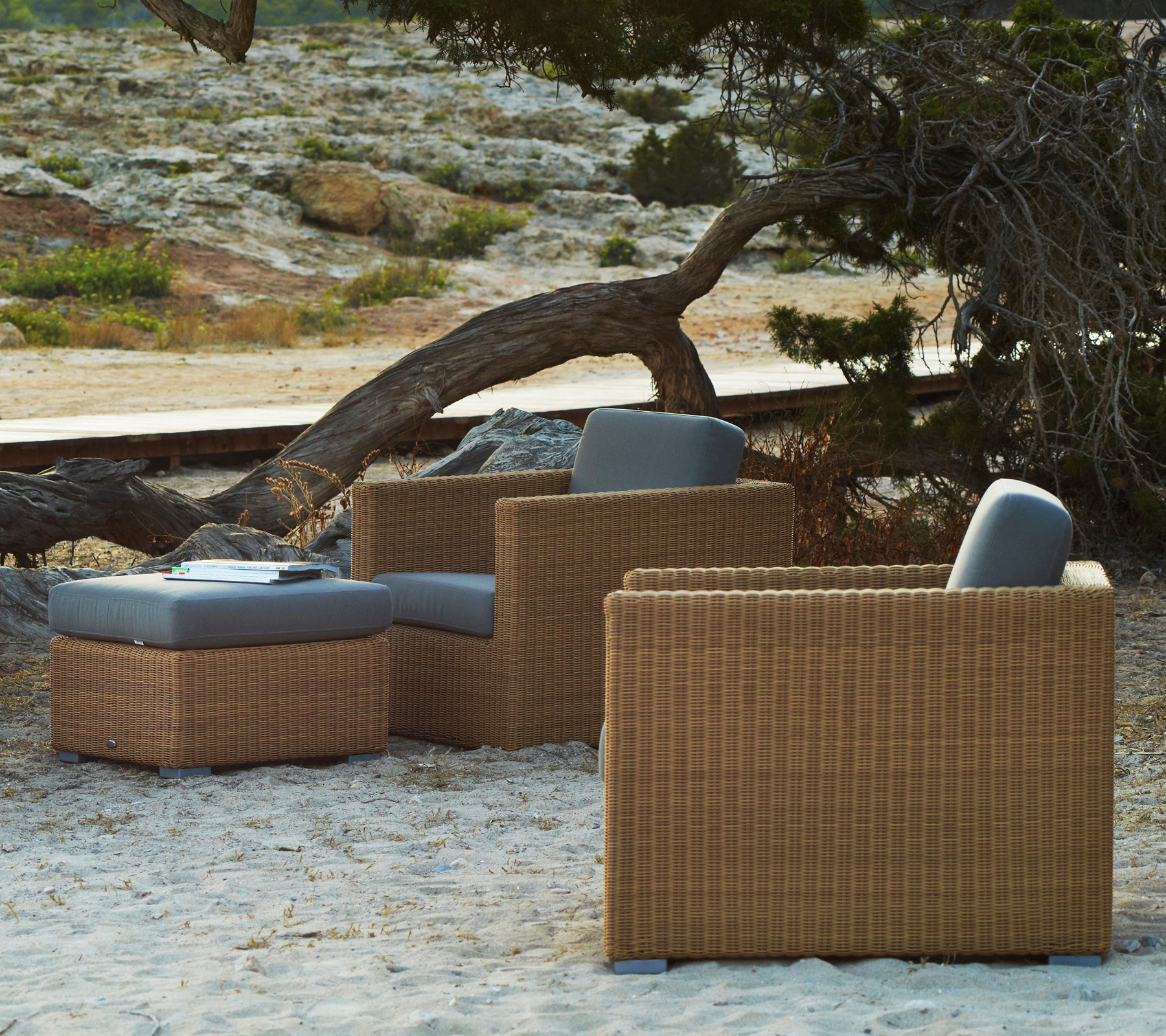 Chaises longues en rotin avec des coussins gris dans un cadre extérieur naturel.