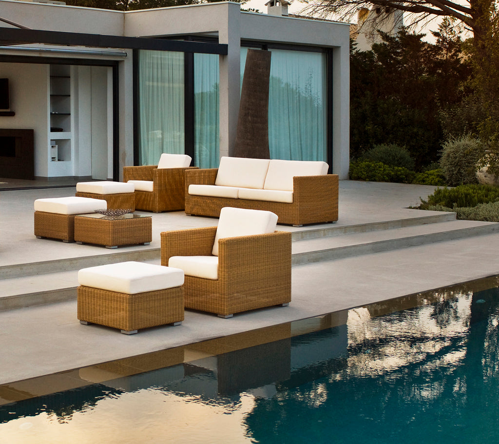 Espace lounge extérieur moderne avec un canapé marron, des chaises et des coussins au bord d'une piscine.