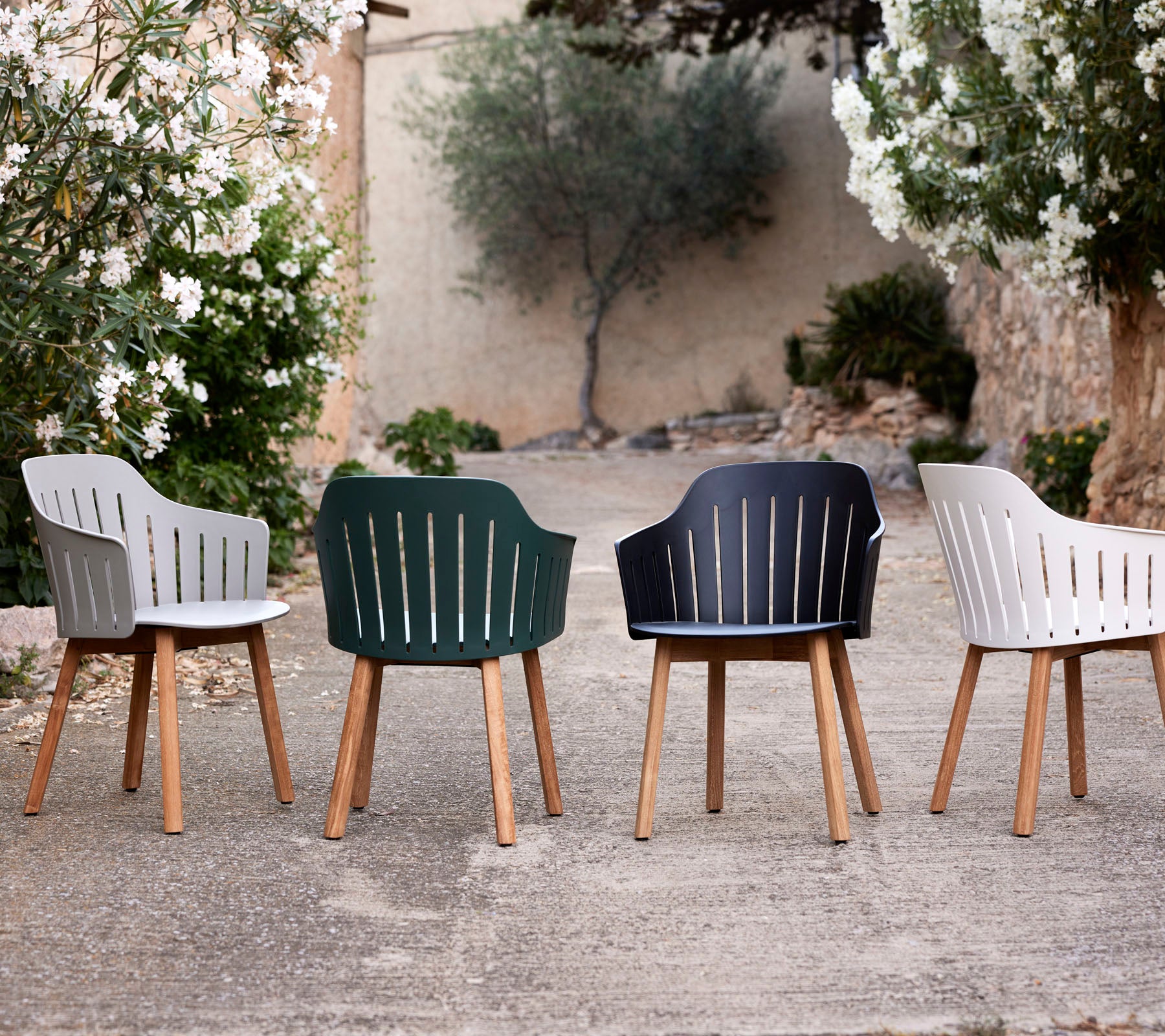 Quatre chaises élégantes de différentes couleurs avec des pieds en bois, présentant un design moderne et des dossiers à lattes au milieu de la verdure.