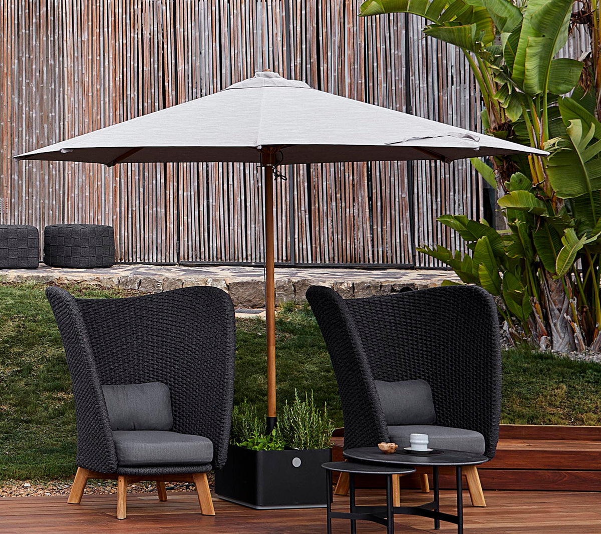 Deux chaises tressées élégantes avec des coussins se trouvent sous un grand parasol, accompagnées d'une petite table, devant un arrière-plan verdoyant.