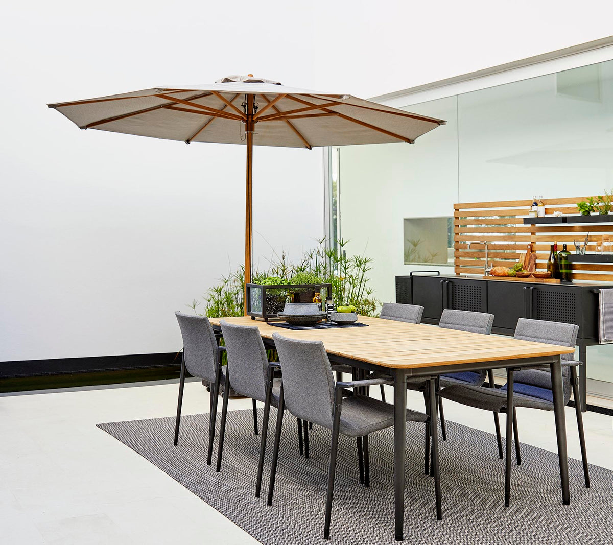 Un ensemble de salle à manger classique avec une table en bois, un parasol et des chaises grises, entouré de plantes décoratives sur un tapis élégant.