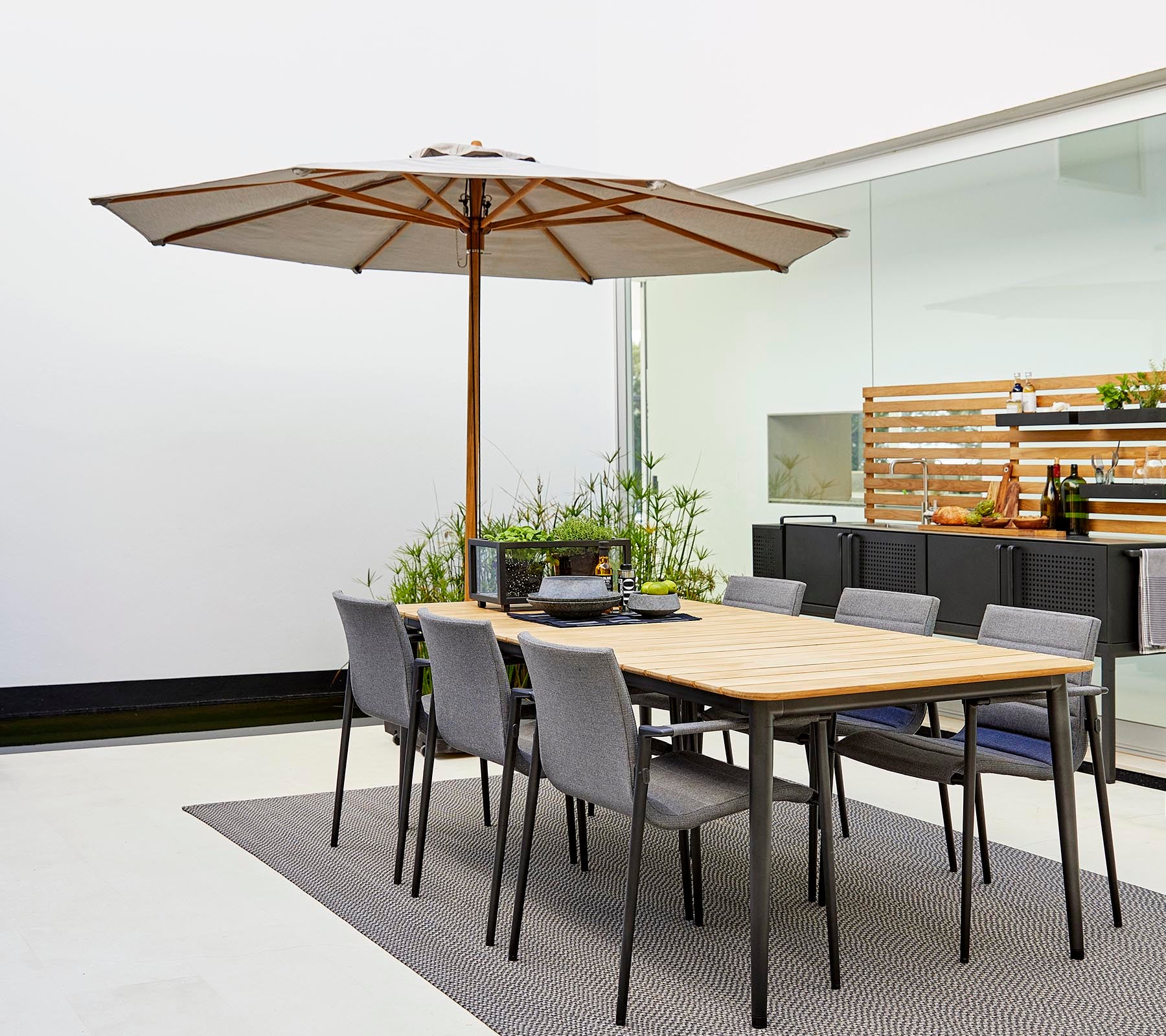 Un ensemble de salle à manger classique avec une table en bois, un parasol et des chaises grises, entouré de plantes décoratives sur un tapis élégant.