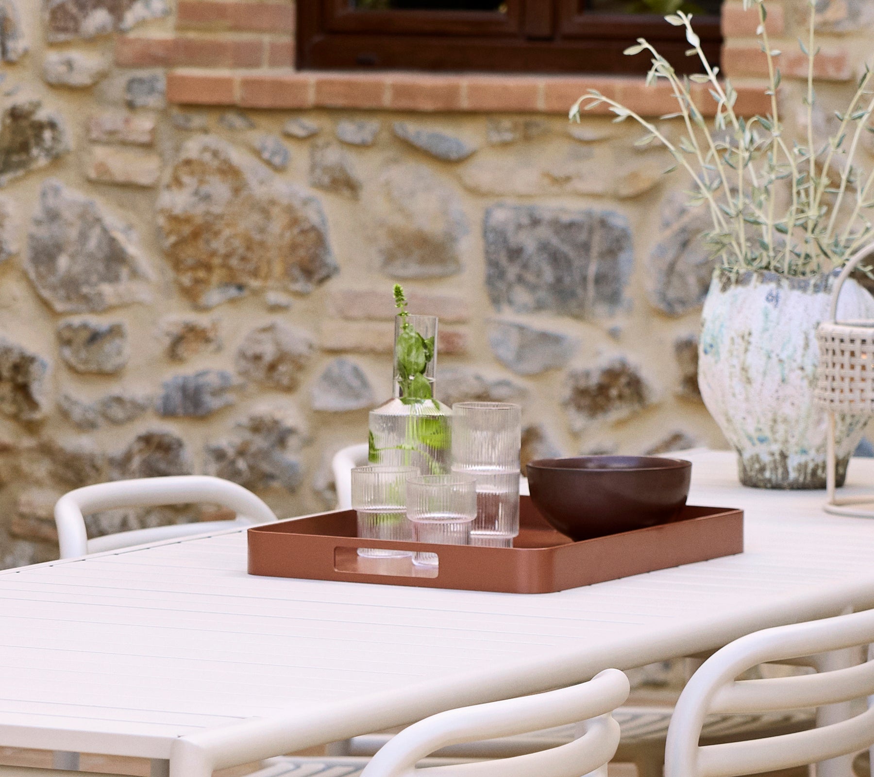 Un plateau de club en rouge désert accueille des verres et un bol, accompagné d'un vase décoratif et de verdure contre un fond en pierre.