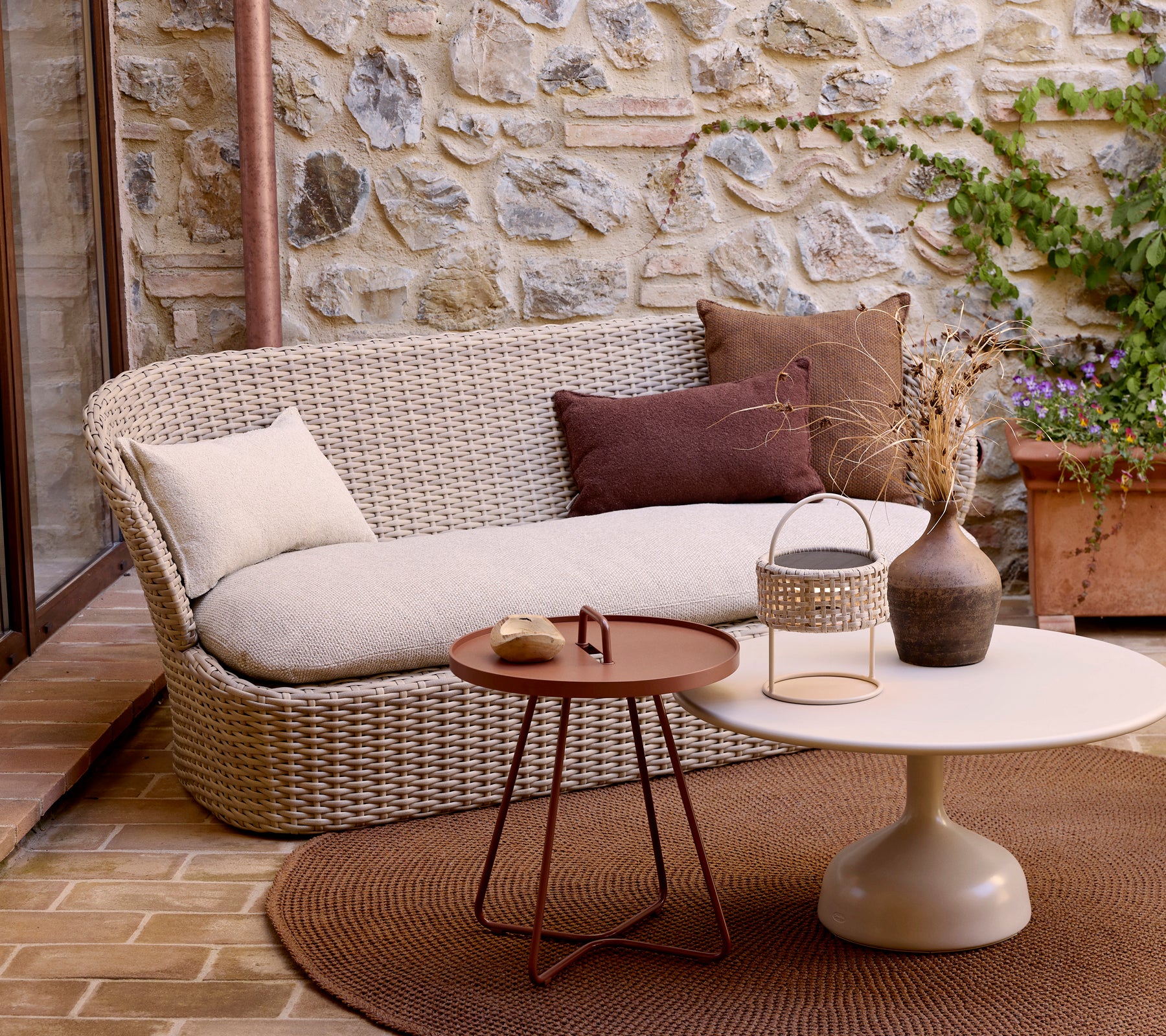 Un canapé moderne pour deux personnes avec des coussins décoratifs, associé à une table basse ronde et des plantes, devant un fond en pierre.