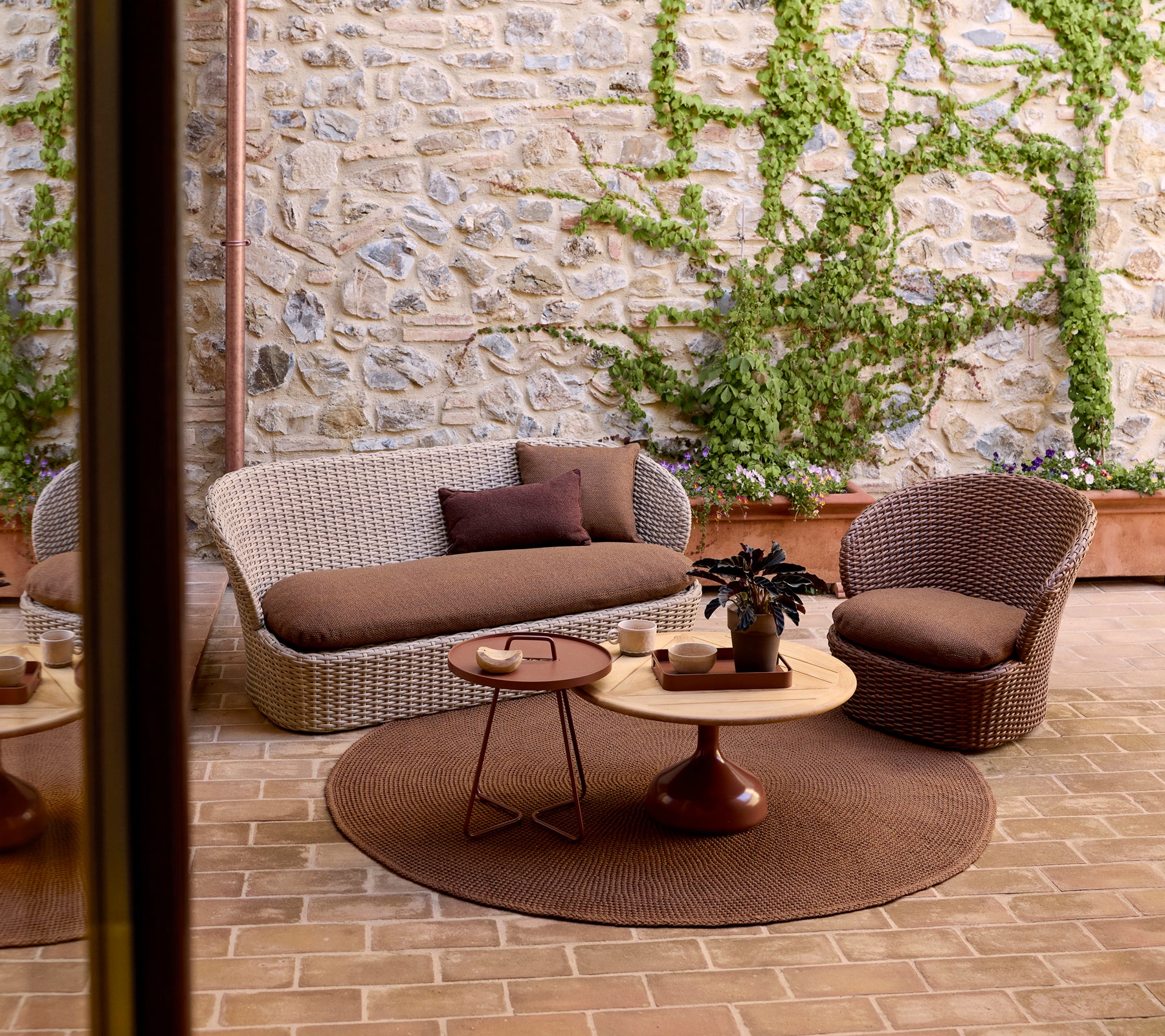 Espace salon extérieur confortable avec un canapé tressé, une chaise, une table d'appoint ronde et de la verdure contre un mur en pierre.