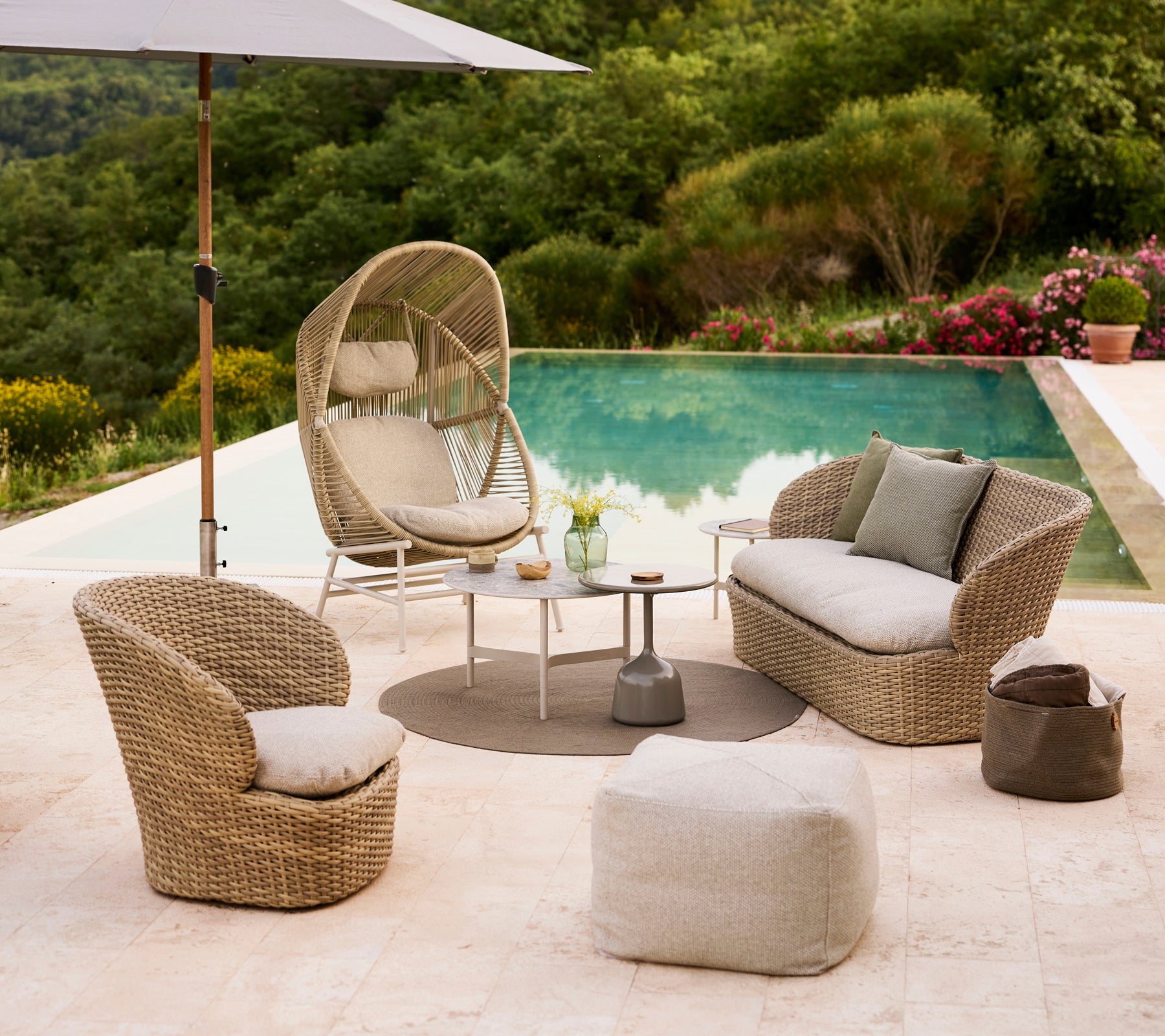 Configuration de salon côtier avec une chaise Hive, deux chaises courbées, un pouf Divine et une petite table près d'une piscine entourée de verdure.
