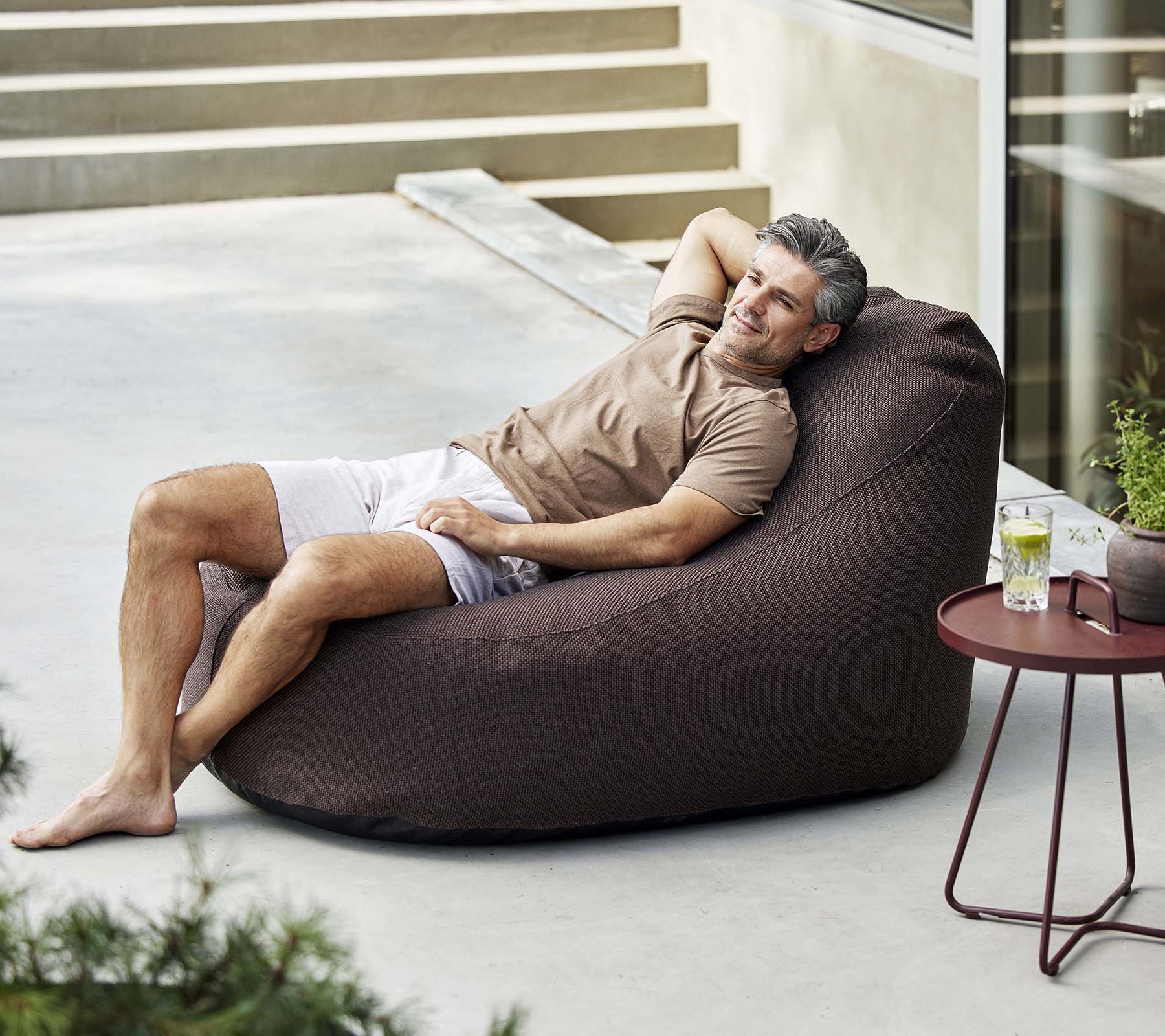Un homme se détend dans un fauteuil confortable, en appréciant une boisson à côté d'une petite table, entouré de verdure.