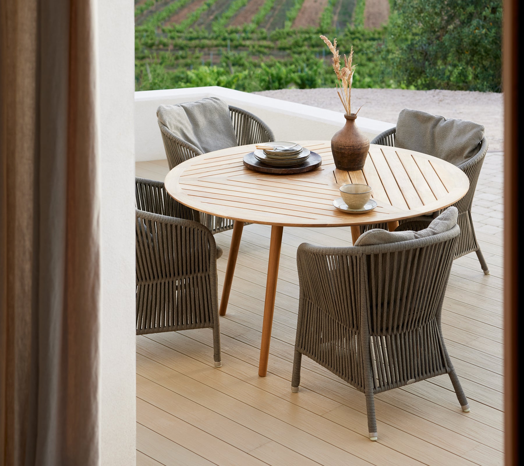 Une table ronde en bois entourée de quatre chaises tressées, avec un vase et un plat décoratif, devant un décor pittoresque.