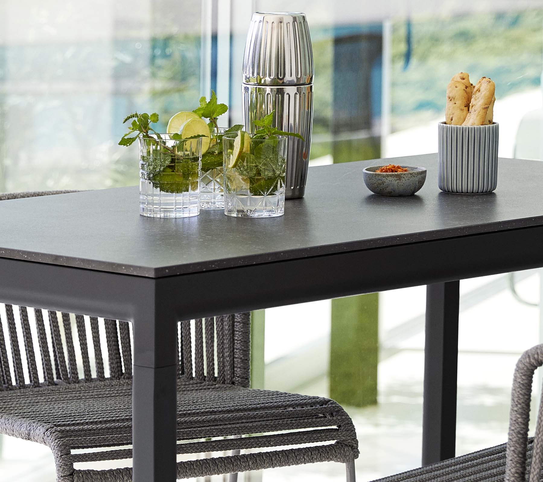 Une table moderne garnie de plantes dans des vases en verre, un contenant décoratif et un plat de collations, entourée de chaises élégantes.