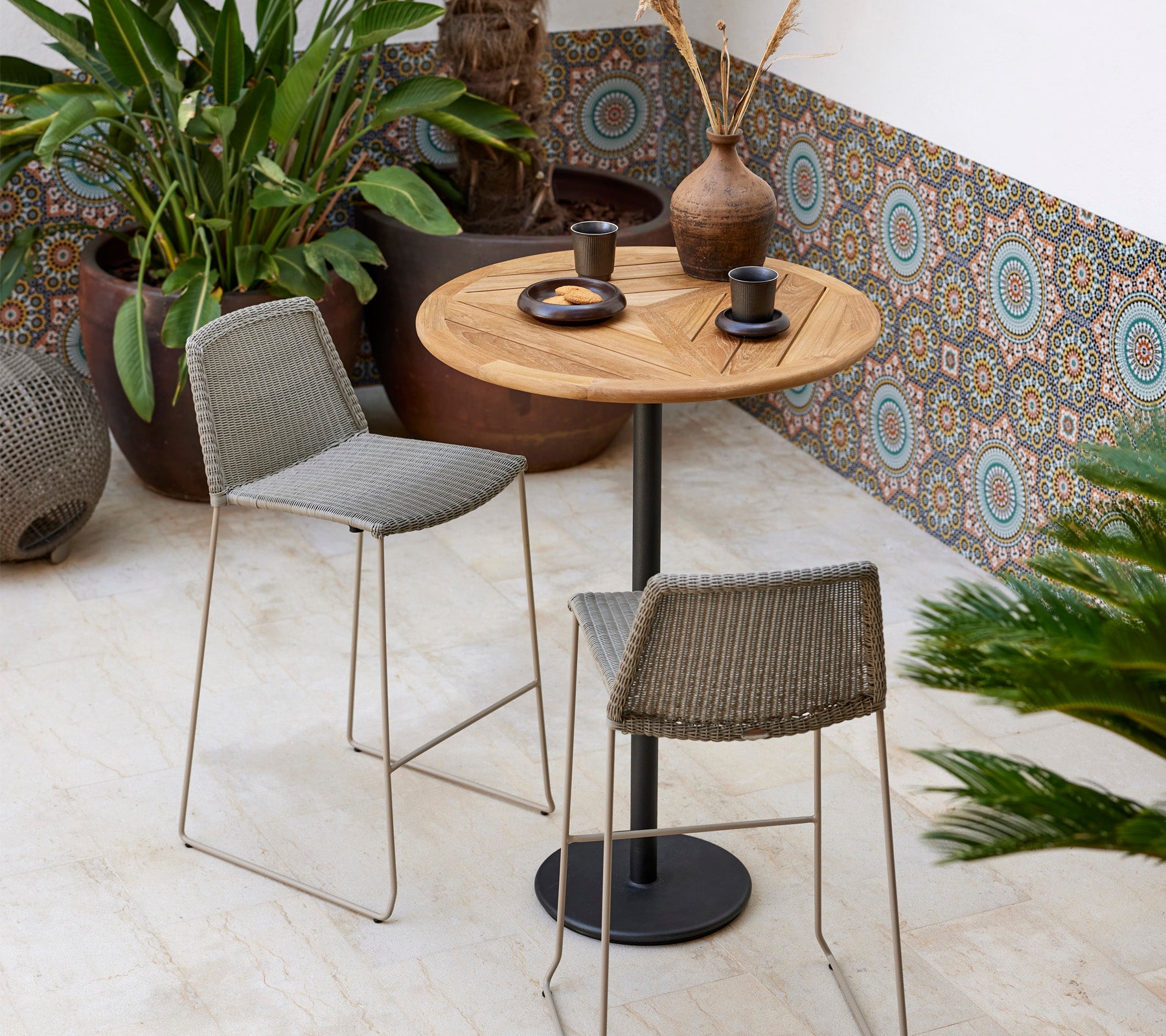 Un ensemble bistro élégant avec une table ronde en bois et deux tabourets tressés, accompagné de plantes décoratives et de vaisselle moderne.