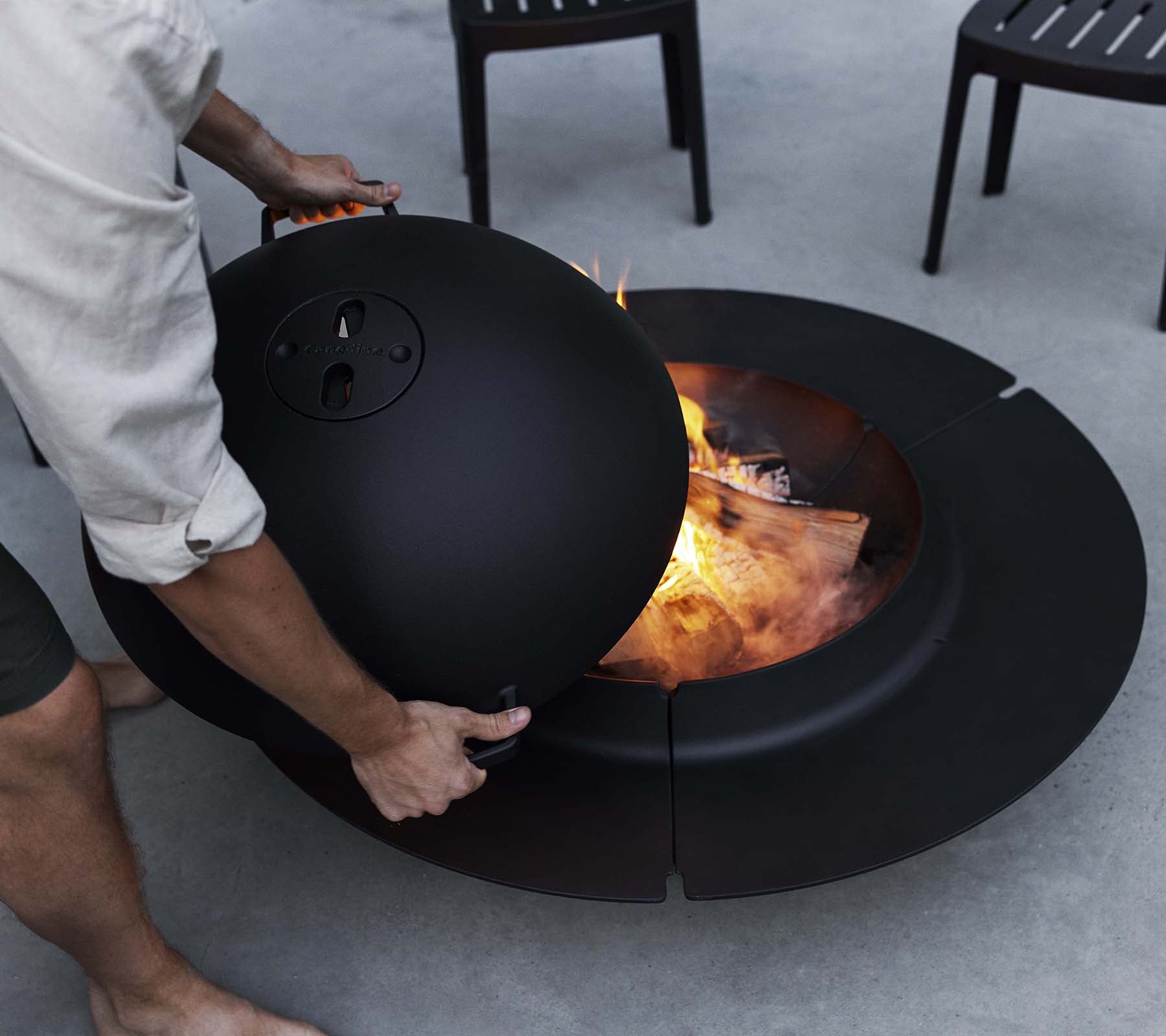 Une personne ajuste un couvercle sur un élégant brasero noir Ember avec des flammes et du bois brûlant visibles à l'intérieur, entouré de chaises.