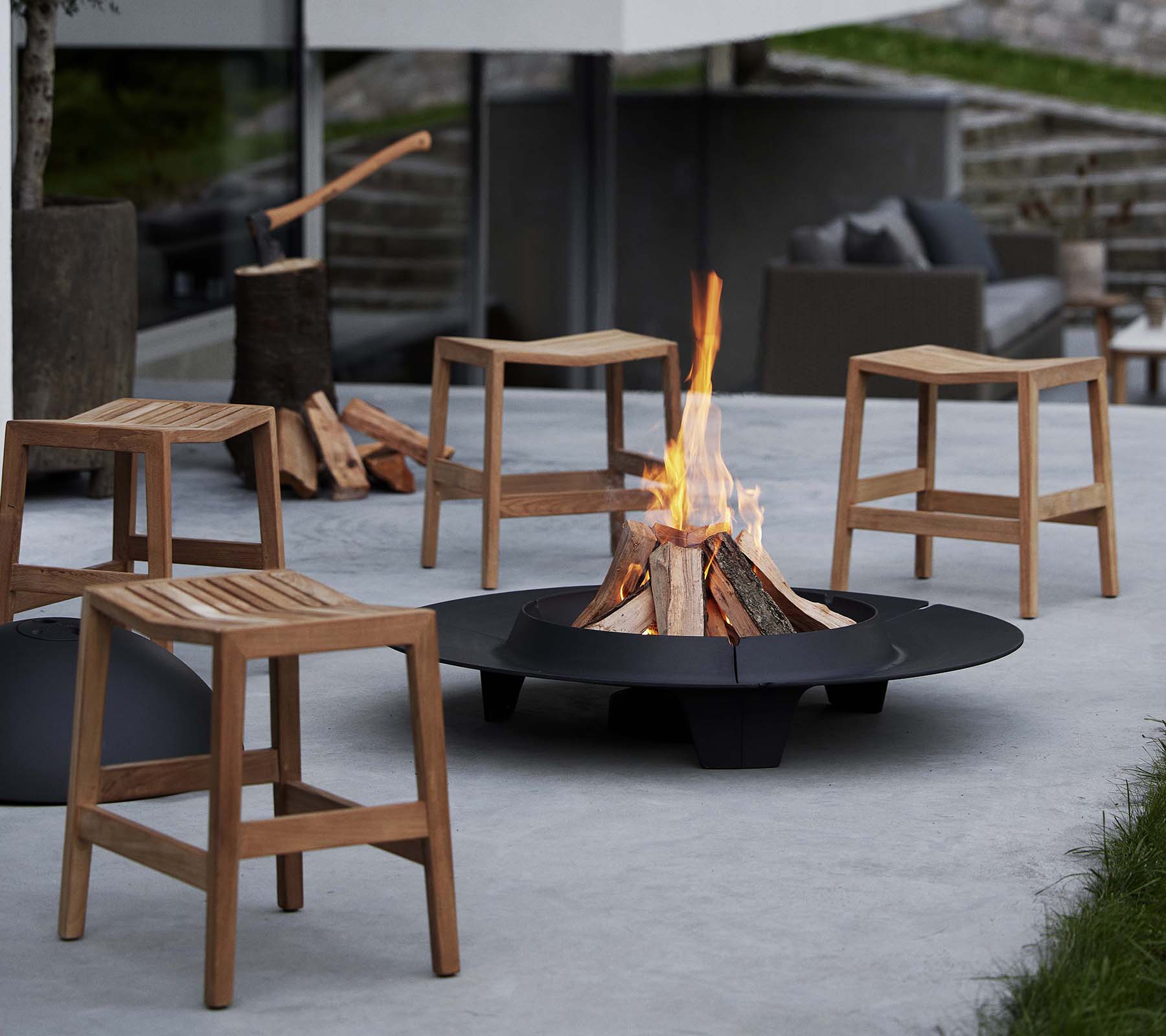 Des tabourets en bois pliants entourent un foyer circulaire de braises, avec un feu flamboyant au centre et des bûches empilées à proximité.