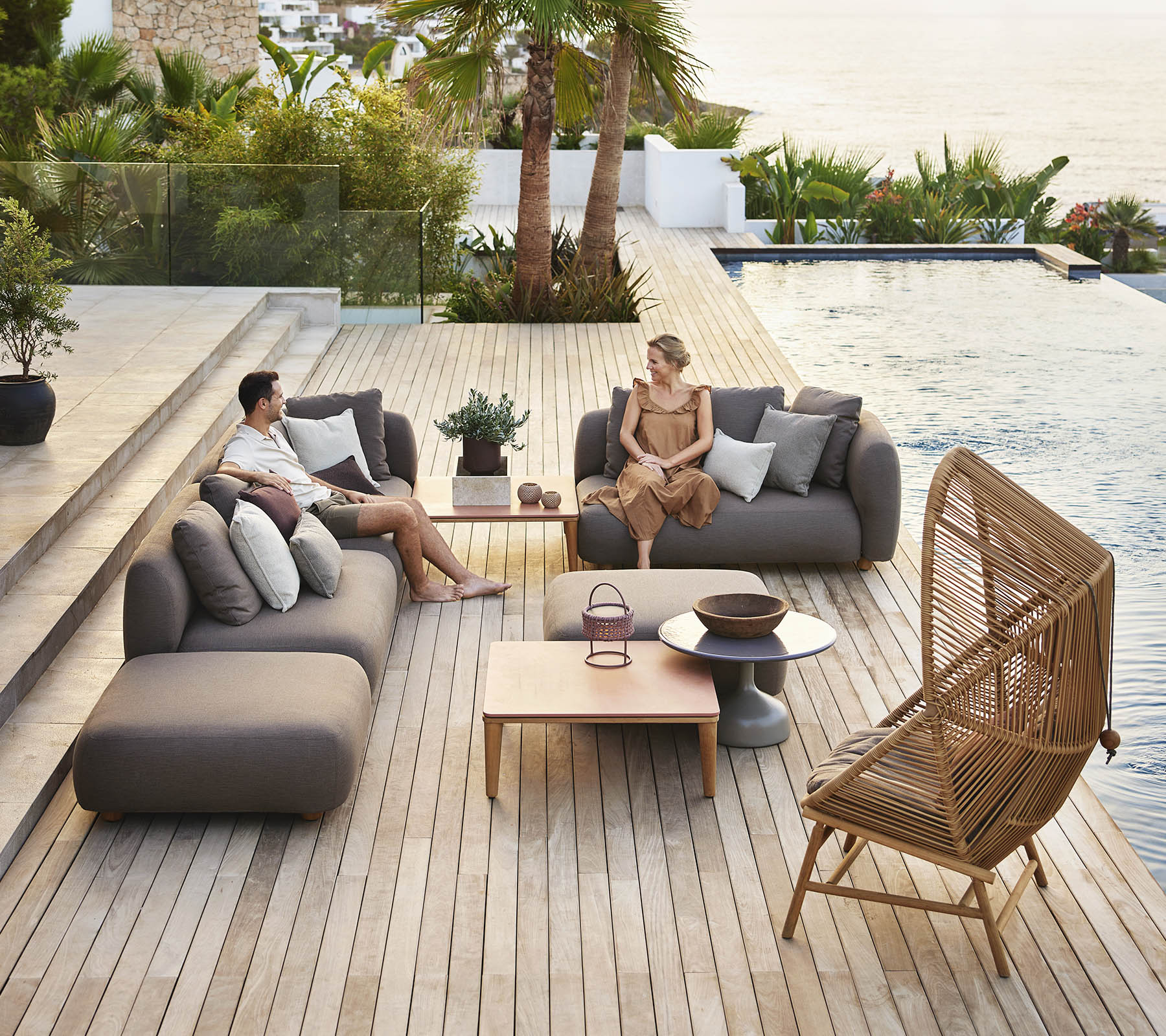 Deux personnes se détendent sur des sièges modernes près d'une piscine, entourées de verdure, avec une table en bois et des plantes décoratives à proximité.