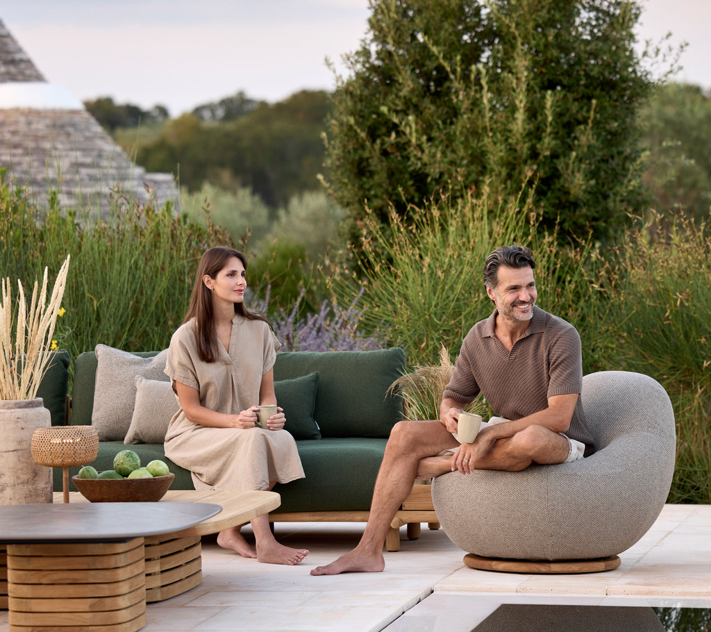 Salon moderne avec canapé vert, chaise ronde grise et table brune entourée de verdure.