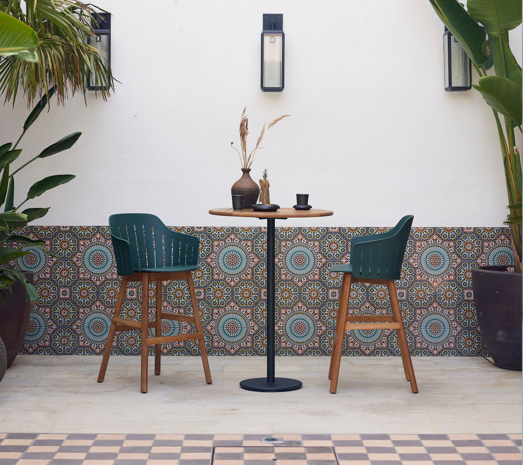 Une table de bar élégante avec deux chaises vertes, décorée d'un vase et d'une tasse, entourée de carreaux décorés et de plantes luxuriantes.