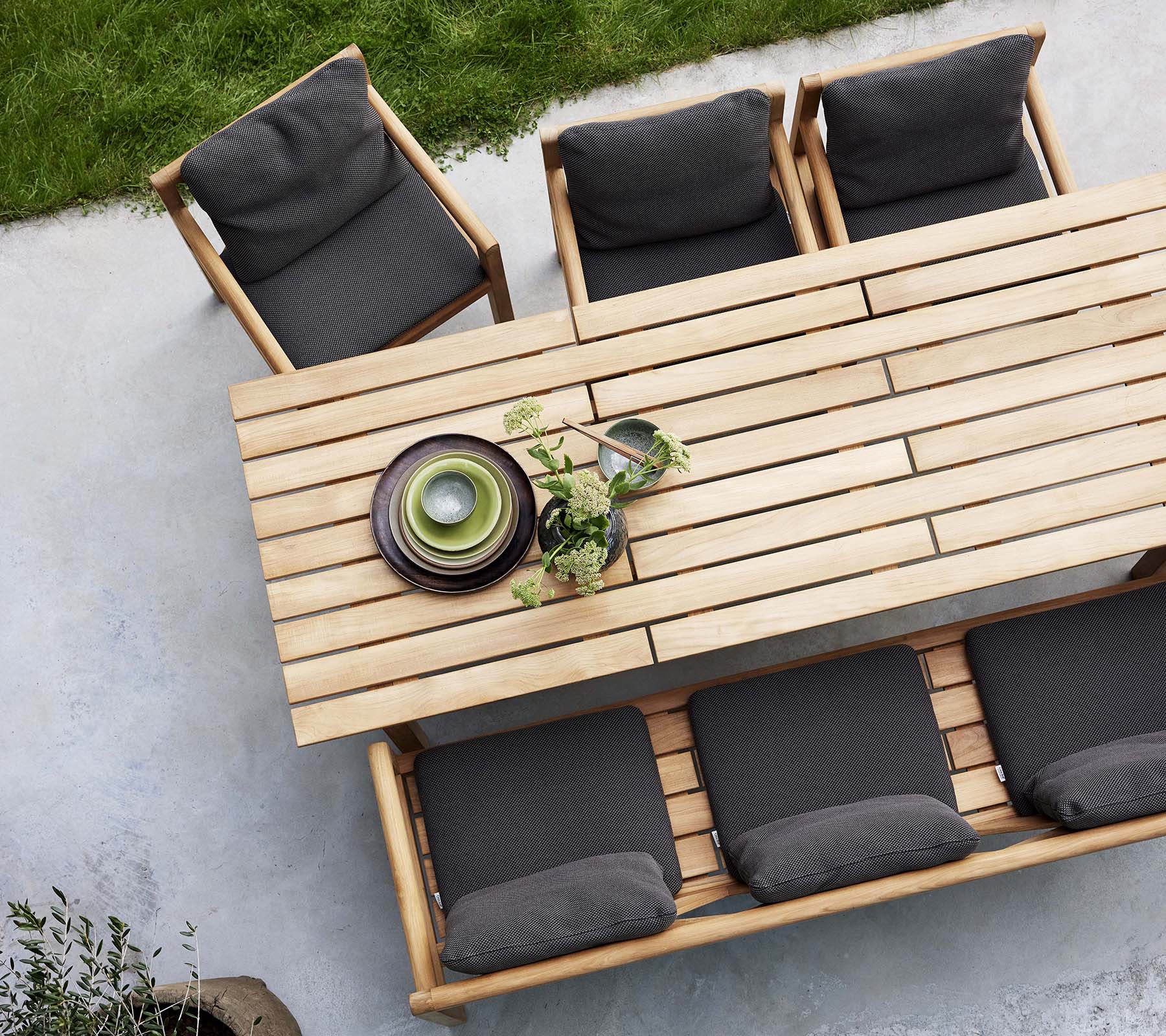 Un ensemble de salle à manger en bois comprenant une table, un banc et des chaises avec des coussins sombres, accentué par des assiettes décoratives et de la verdure.
