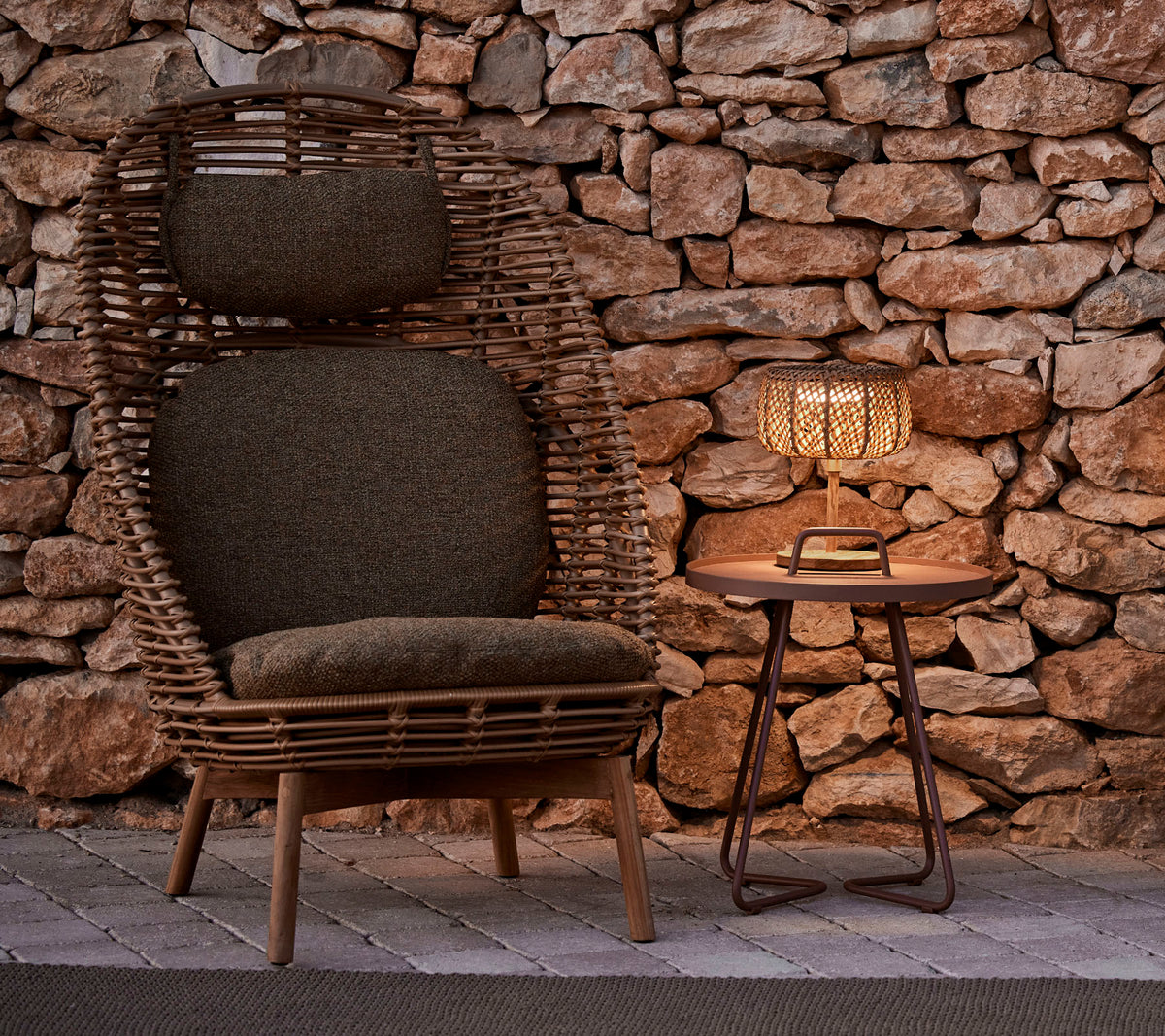 Une chaise tissée élégante avec des coussins à côté d'une table en bois et d'une lampe lumineuse, sur fond de pierre rustique.