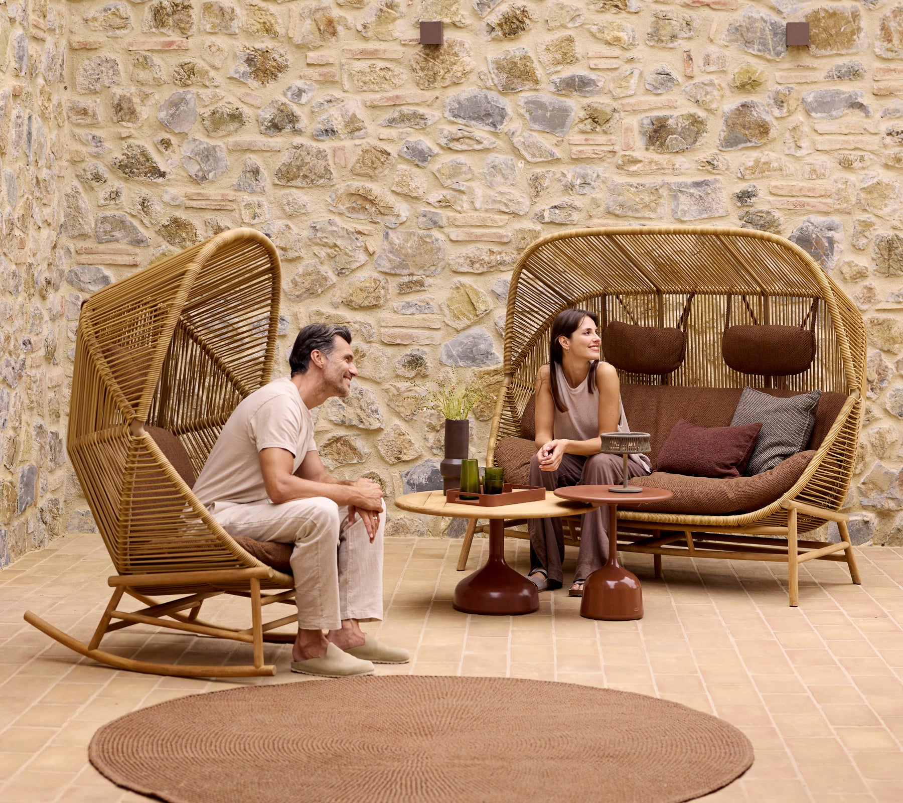 Deux personnes se détendent dans des fauteuils à bascule Hive élégants et un canapé Hive, entourés de tables basses Glaze et d'un doux tapis en tricot.