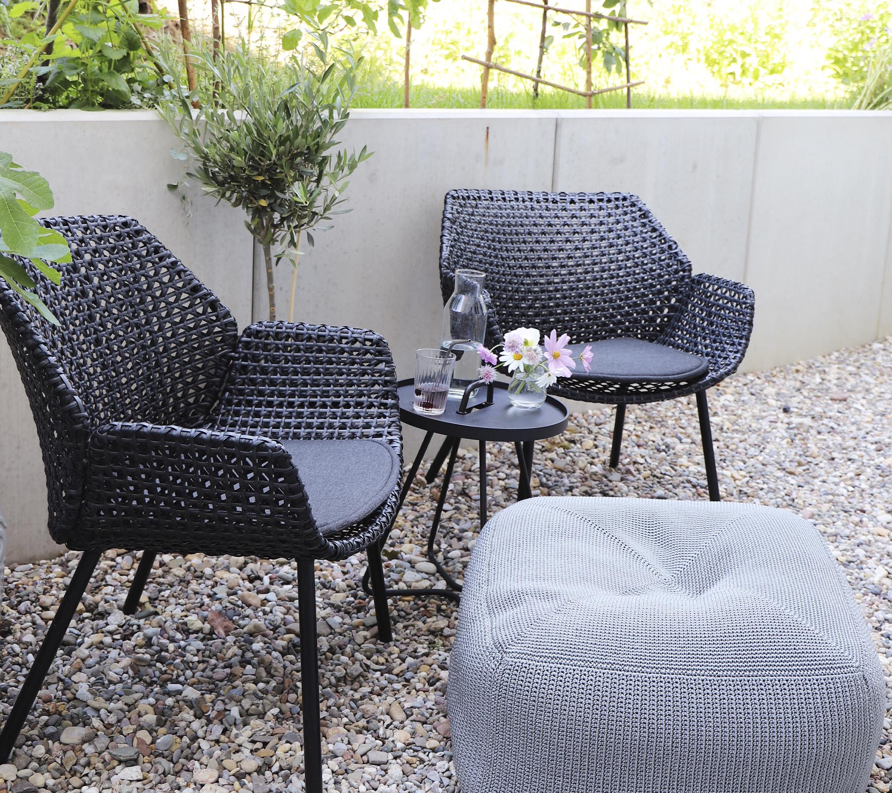 Deux chaises noires élégantes associées à une petite table, ornées d'un vase de fleurs et d'un pouf confortable sur un sol en gravier.