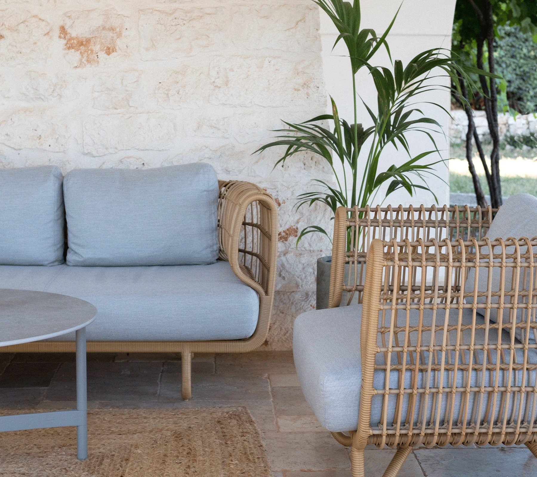 Espace de détente cosy avec un canapé gris clair, des fauteuils en osier, une table ronde et de la verdure en arrière-plan. Idéal pour se détendre.