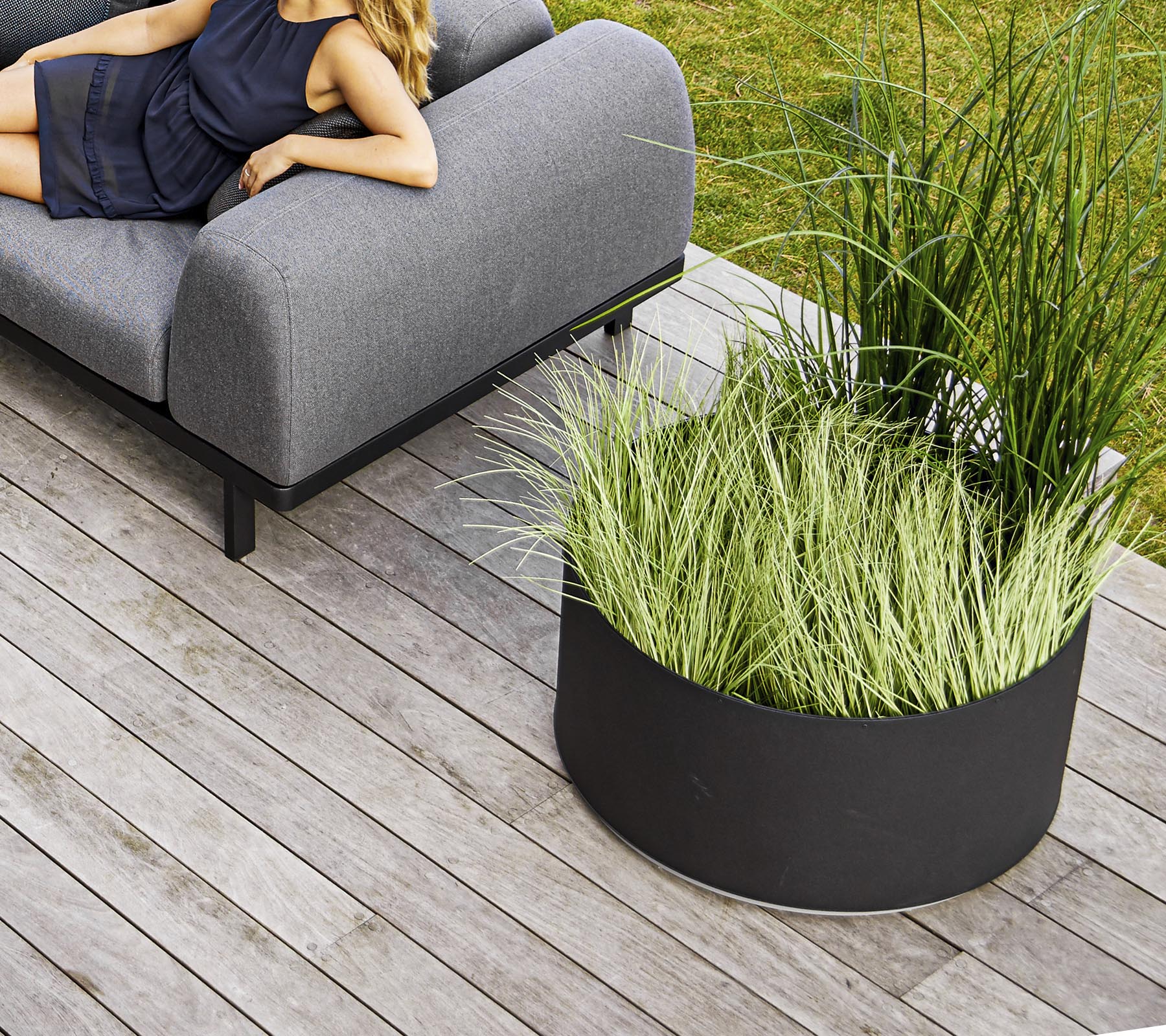 Un canapé gris élégant à côté d'un pot circulaire noir rempli de hautes herbes vertes, placé sur une terrasse en bois.