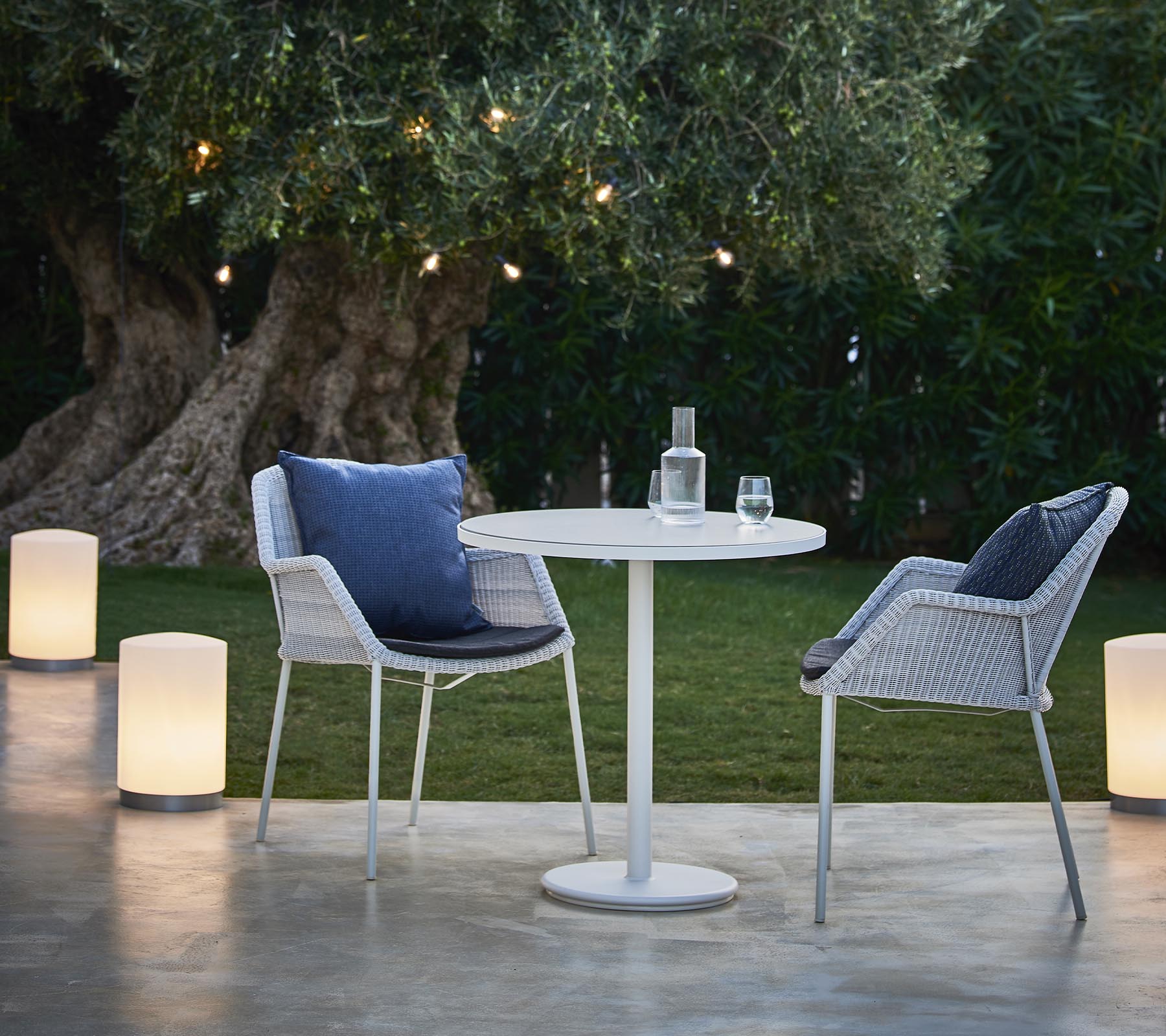 Un coin salon confortable avec deux chaises modernes, une table ronde et des lampes élégantes, entouré de verdure et d'un éclairage chaleureux.