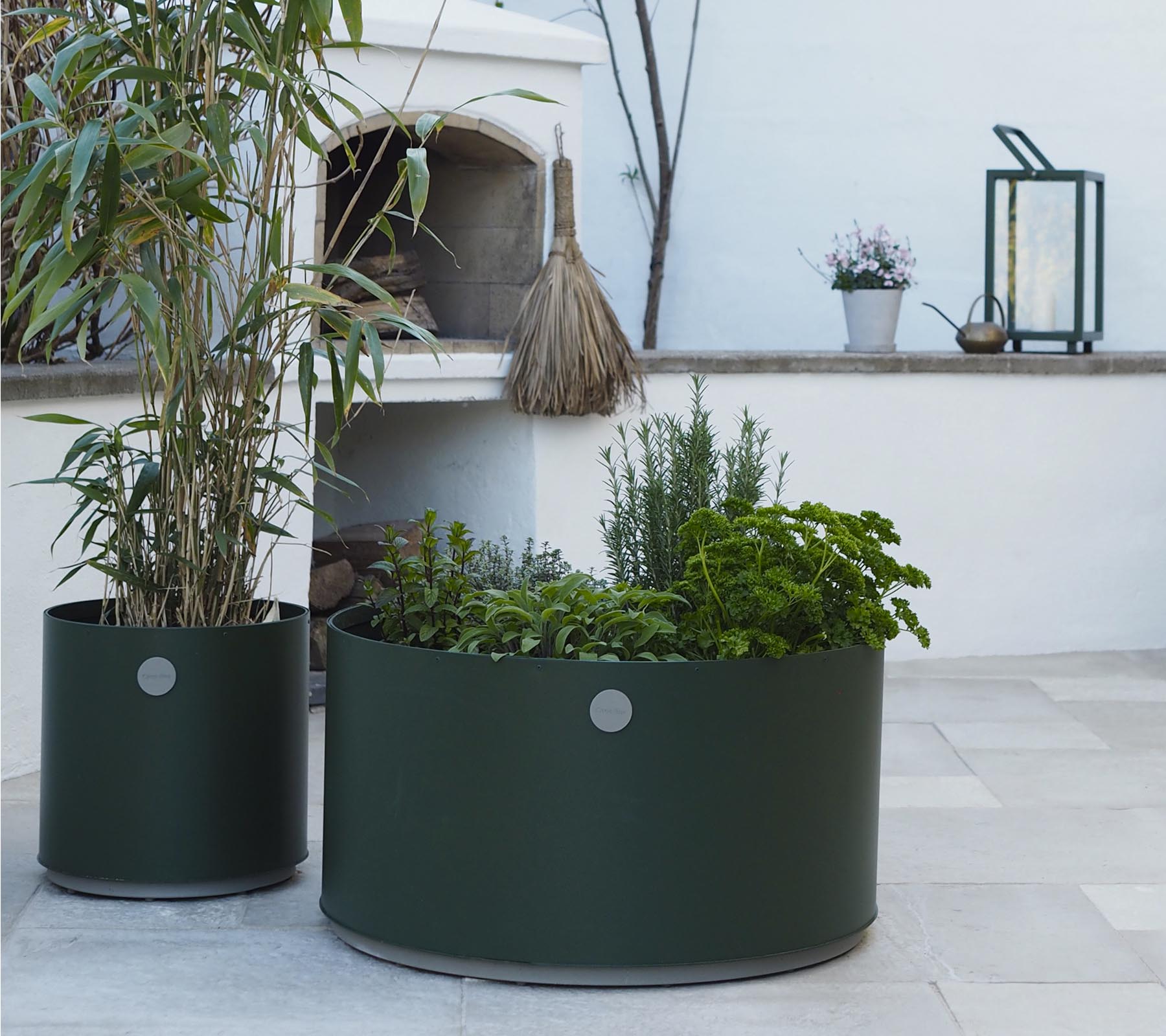 Deux jardinières rondes en vert foncé, l'une présentant des plantes hautes et l'autre remplie de diverses herbes, reposent sur une surface en pierre claire.