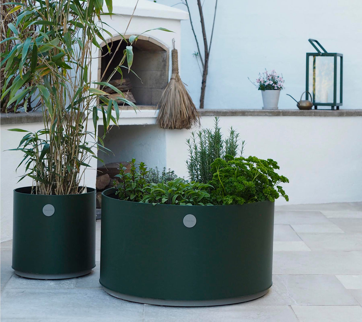 Deux jardinières rondes en vert foncé, l'une présentant des plantes hautes et l'autre remplie de diverses herbes, reposent sur une surface en pierre claire.