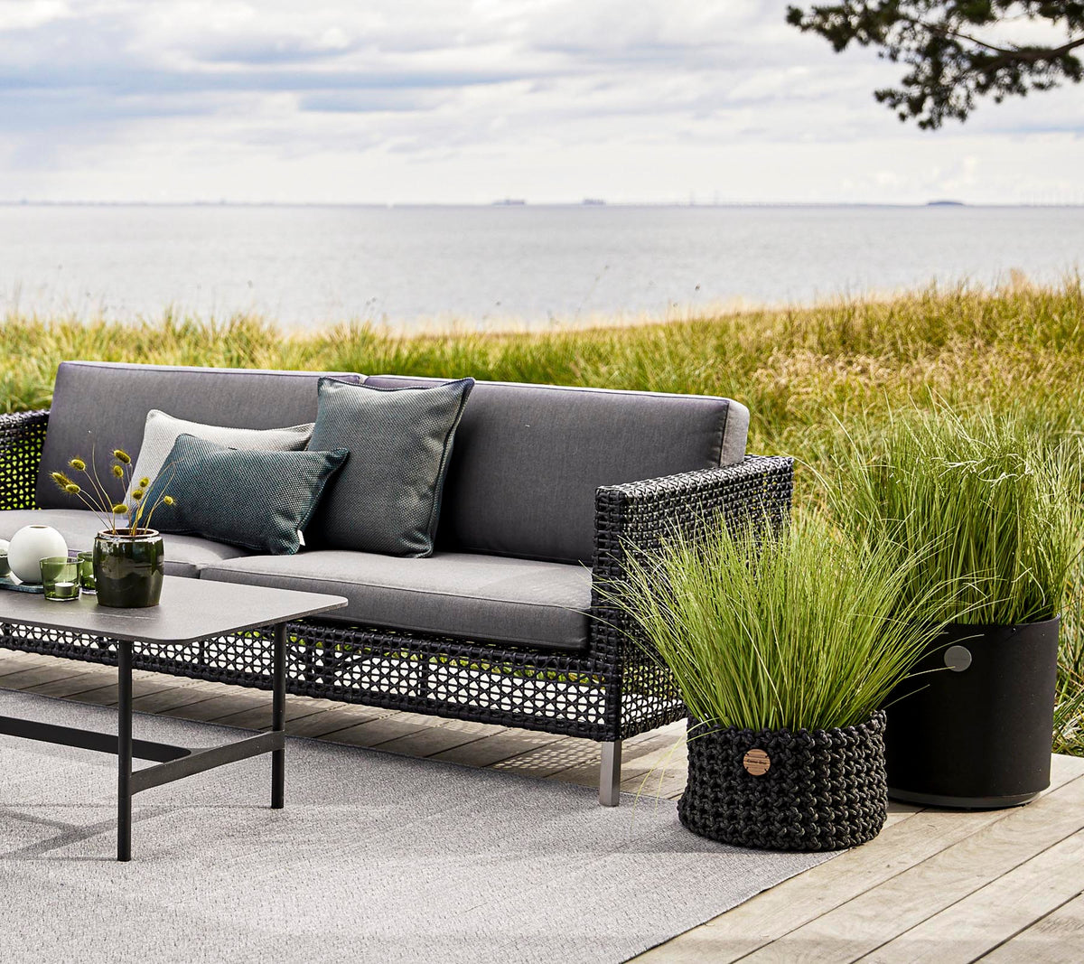 Mobilier d'extérieur élégant avec un canapé gris, des coussins décoratifs et une table, complété par des jardinières décoratives en herbe et un décor serein.