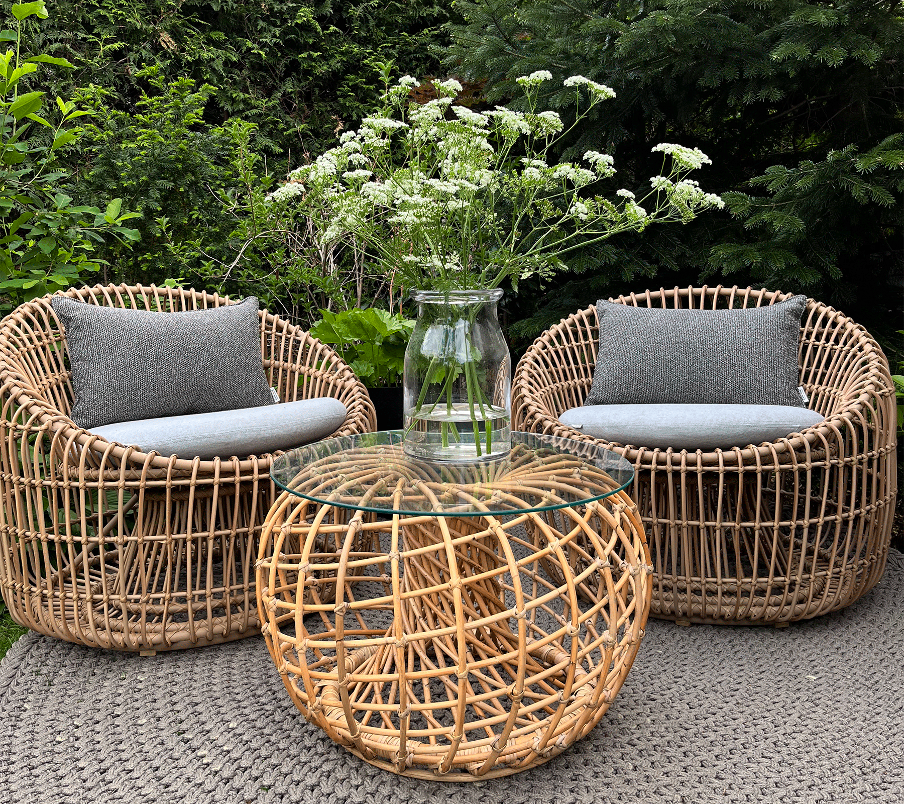 Deux chaises tissées avec des coussins gris entourent une table basse ronde tissée surmontée d'un vase transparent contenant des fleurs blanches.