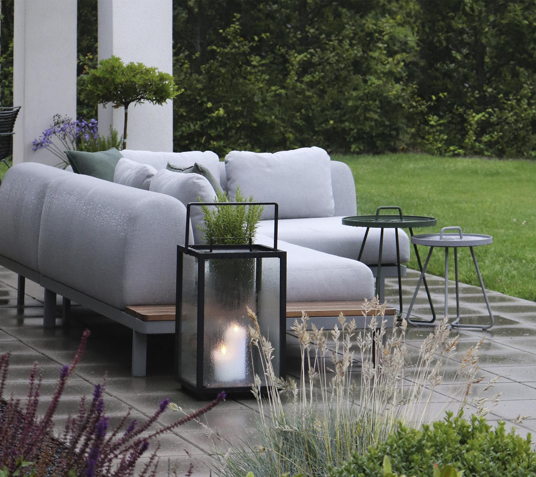 Agencement extérieur élégant avec des coussins moelleux, accompagné de petites tables d'appoint et d'une lanterne, entouré de verdure.