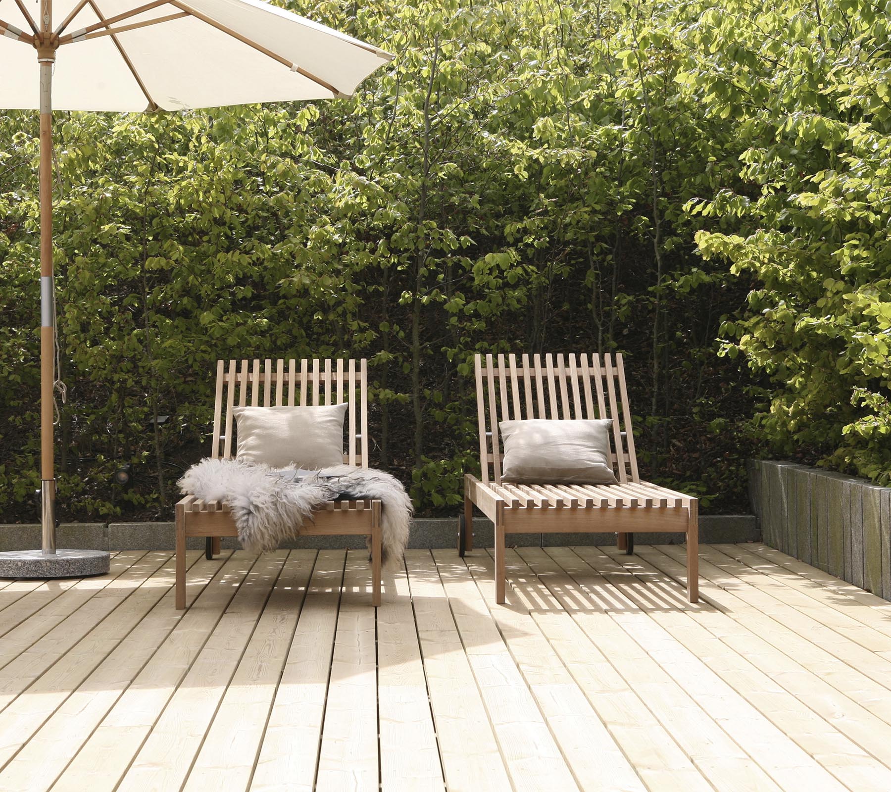 Deux chaises longues en bois élégantes avec coussins, positionnées sur une terrasse en bois, entourées de verdure et d'un grand parasol à proximité.