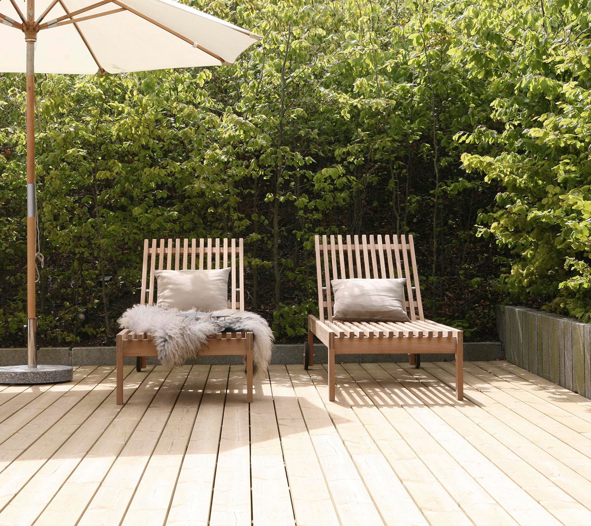 Deux chaises longues en bois élégantes avec coussins, positionnées sur une terrasse en bois, entourées de verdure et d'un grand parasol à proximité.