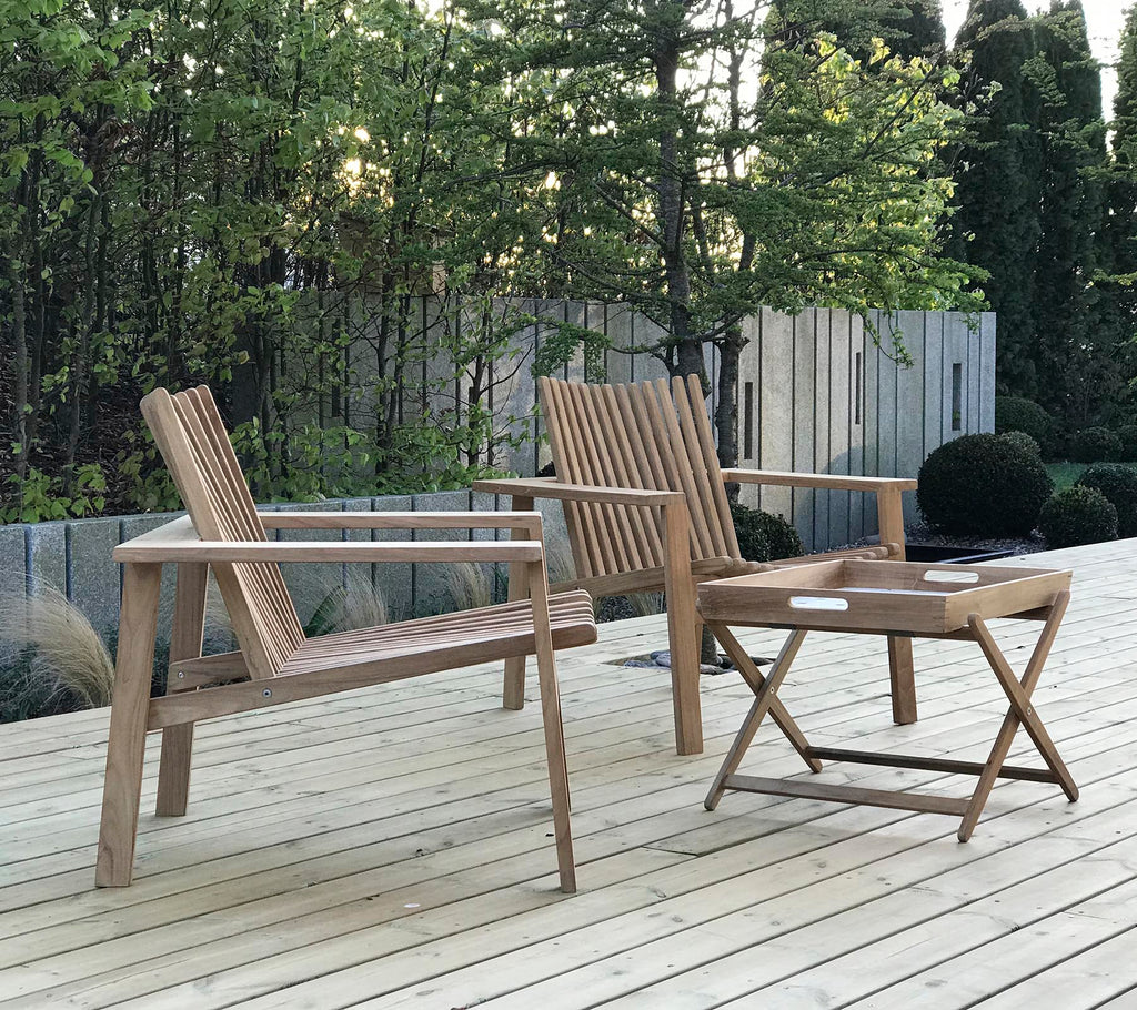Deux chaises en bois et une petite table pliante, placées devant un arrière-plan de verdure et de panneaux en bois. Assises extérieures naturelles et accueillantes.