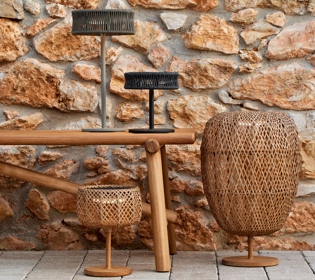 Trois lampes décoratives de différentes formes et tailles sur une table en bois, contre un mur en pierre texturé.