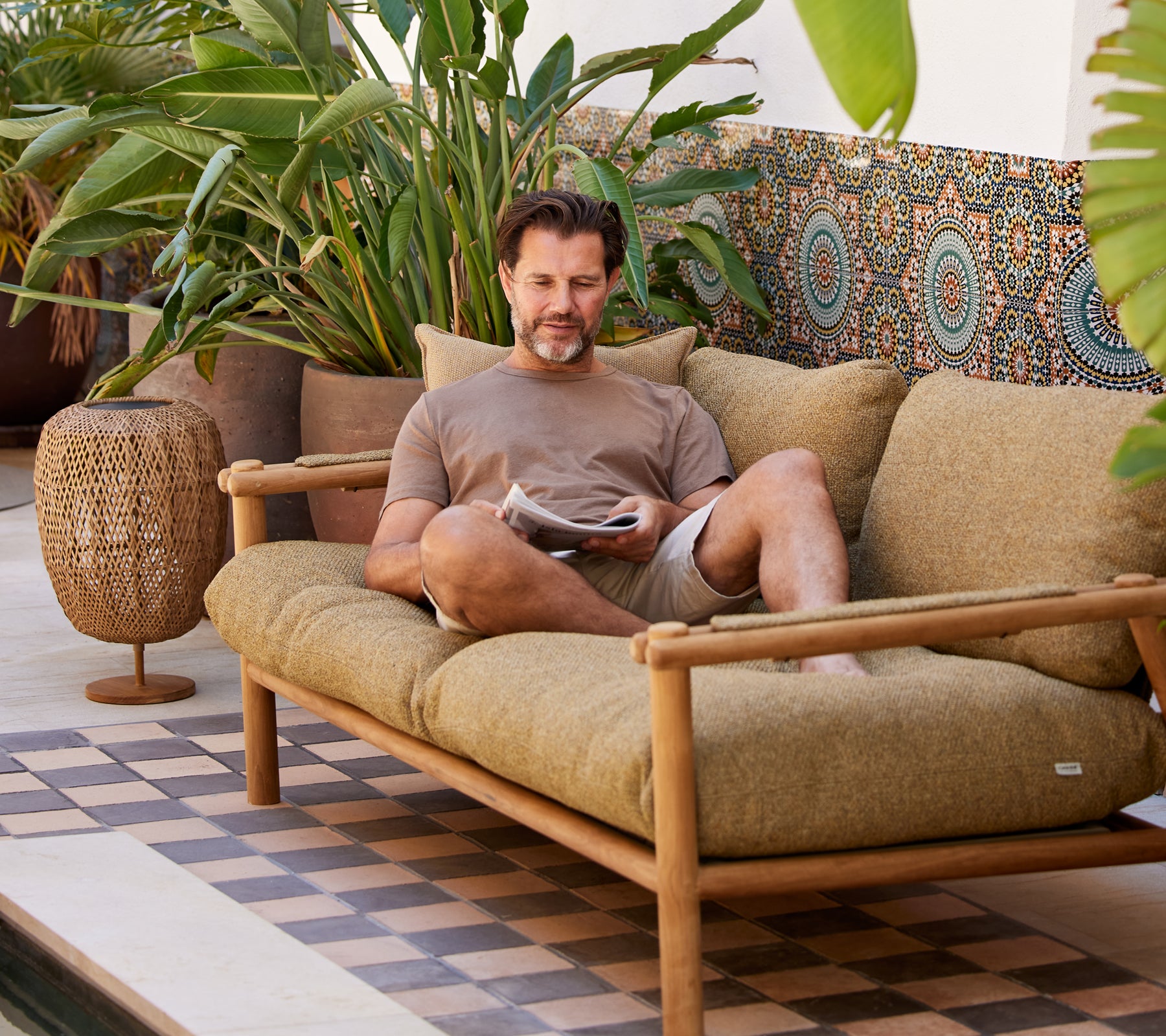 Un homme se détend sur un canapé confortable, lisant un magazine, entouré d'une verdure luxuriante et de motifs décoratifs.