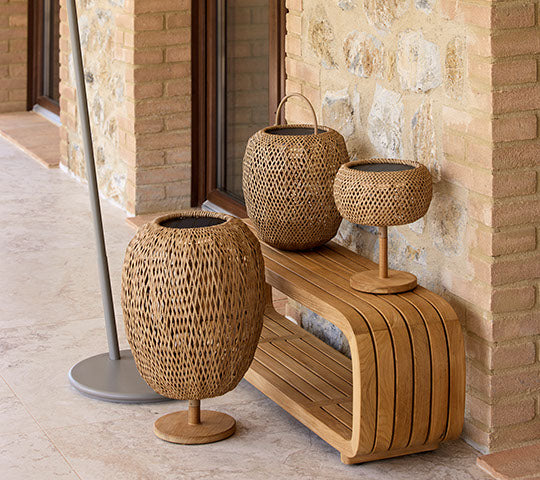 Trois lampes en osier de hauteurs variées disposées sur un banc en bois, complétées par un support élégant. Des matériaux naturels créent une ambiance chaleureuse.