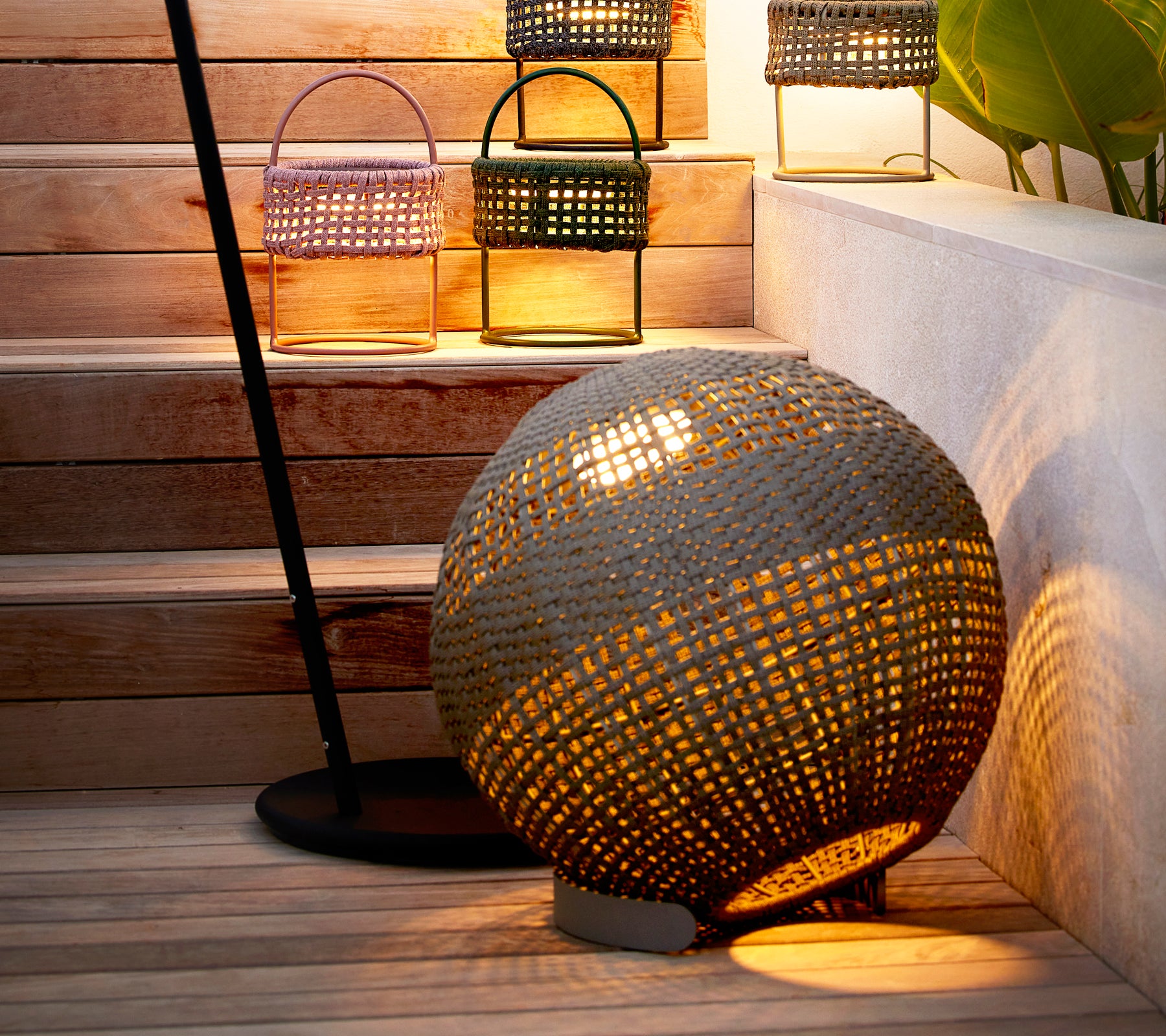 Lampe ronde illusion taupe au design tissé, projetant une chaude lueur à côté de lanternes élégantes sur un escalier.