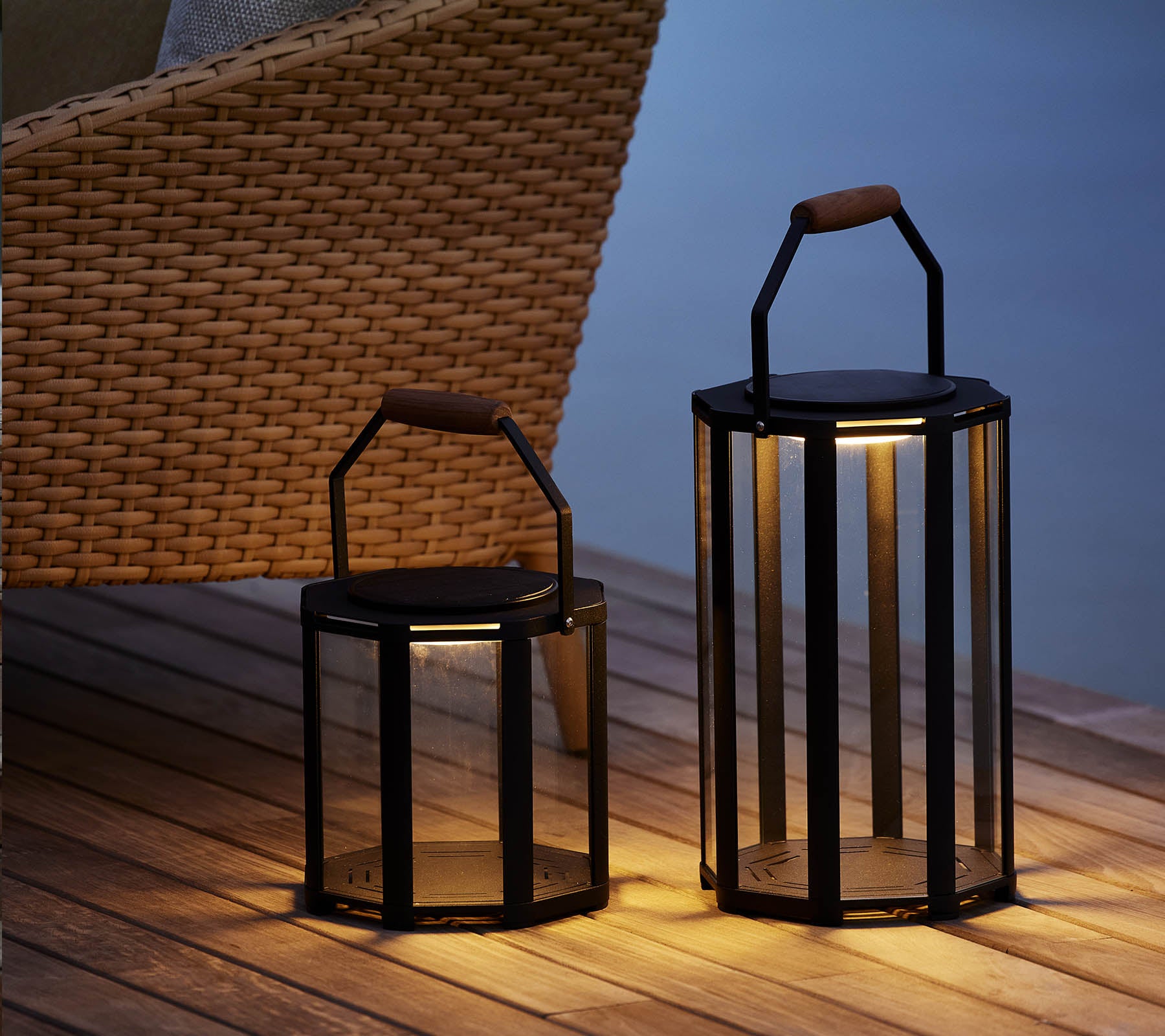 Deux lanternes Lightluxe élégantes avec des panneaux en verre et des poignées, émettant une lueur chaleureuse, placées à côté d'une chaise tissée.
