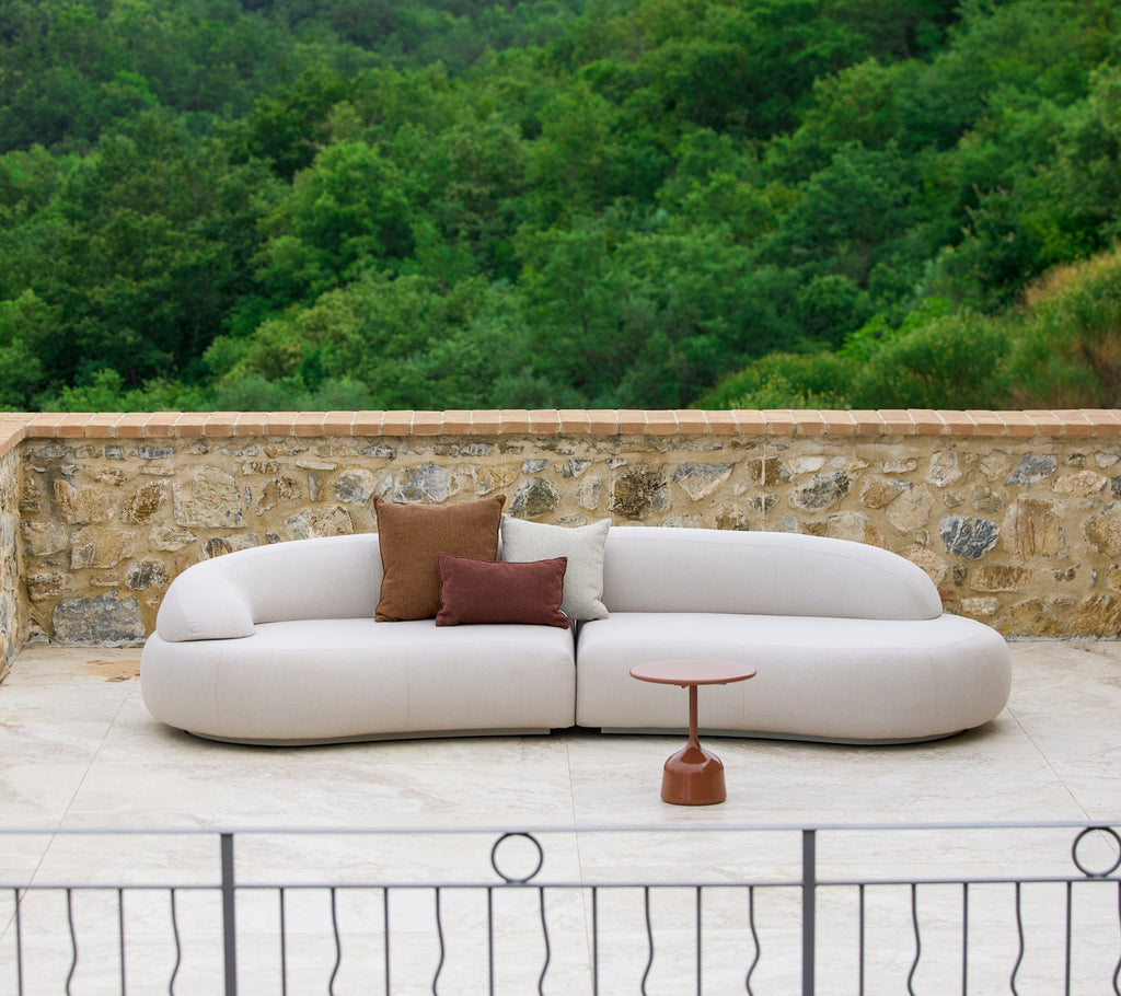 Un canapé moderne et courbe avec un revêtement neutre et des coussins décoratifs, complété par une table d'appoint élégante dans un cadre naturel.
