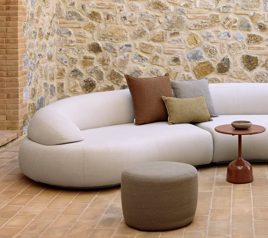 Canapé courbé avec coussins décoratifs, un repose-pieds circulaire et une table d'appoint glazée sur un sol texturé avec un mur en pierre en fond.