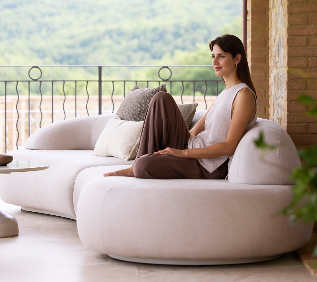 Une femme est assise confortablement sur un canapé moderne et épuré, accompagnée de coussins décoratifs, appréciant une vue sereine sur la nature.