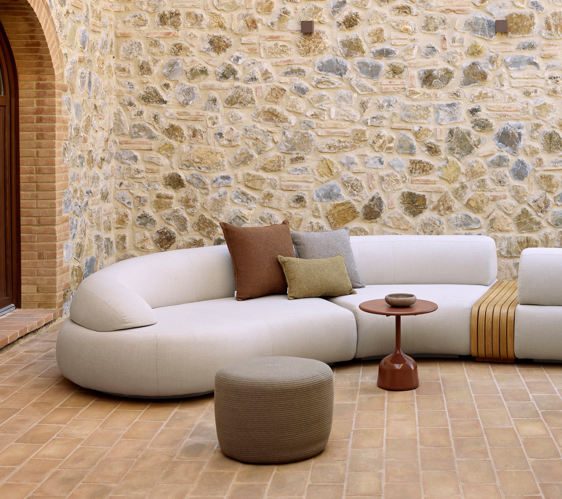 Zone de détente avec un canapé courbé, une table basse Glaze et un pouf rond contre un mur en pierre texturé.