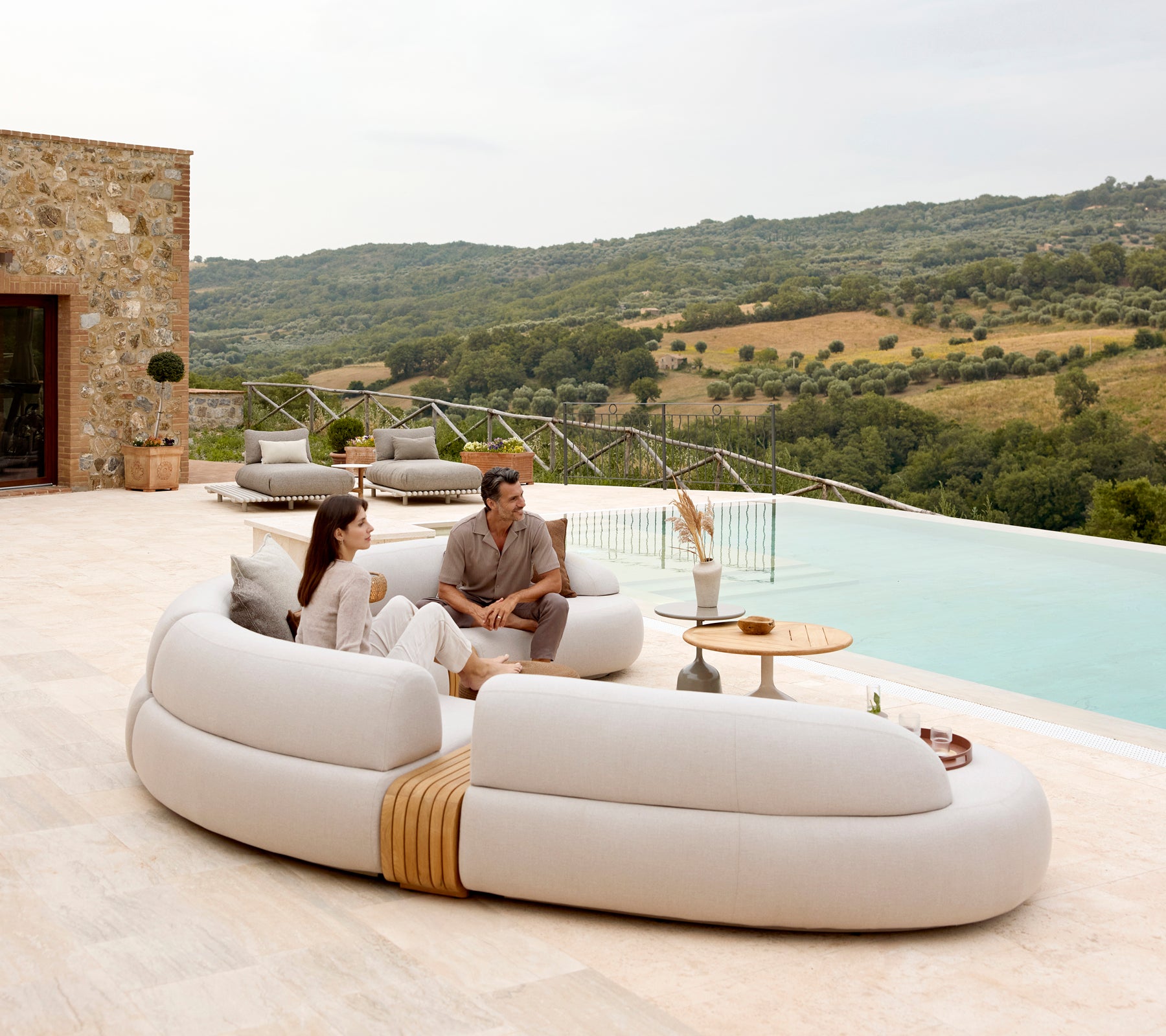 Un couple se détend sur un canapé moderne et courbé, à côté d'une table basse élégante, avec une vue pittoresque sur des collines vallonnées à l'arrière-plan.