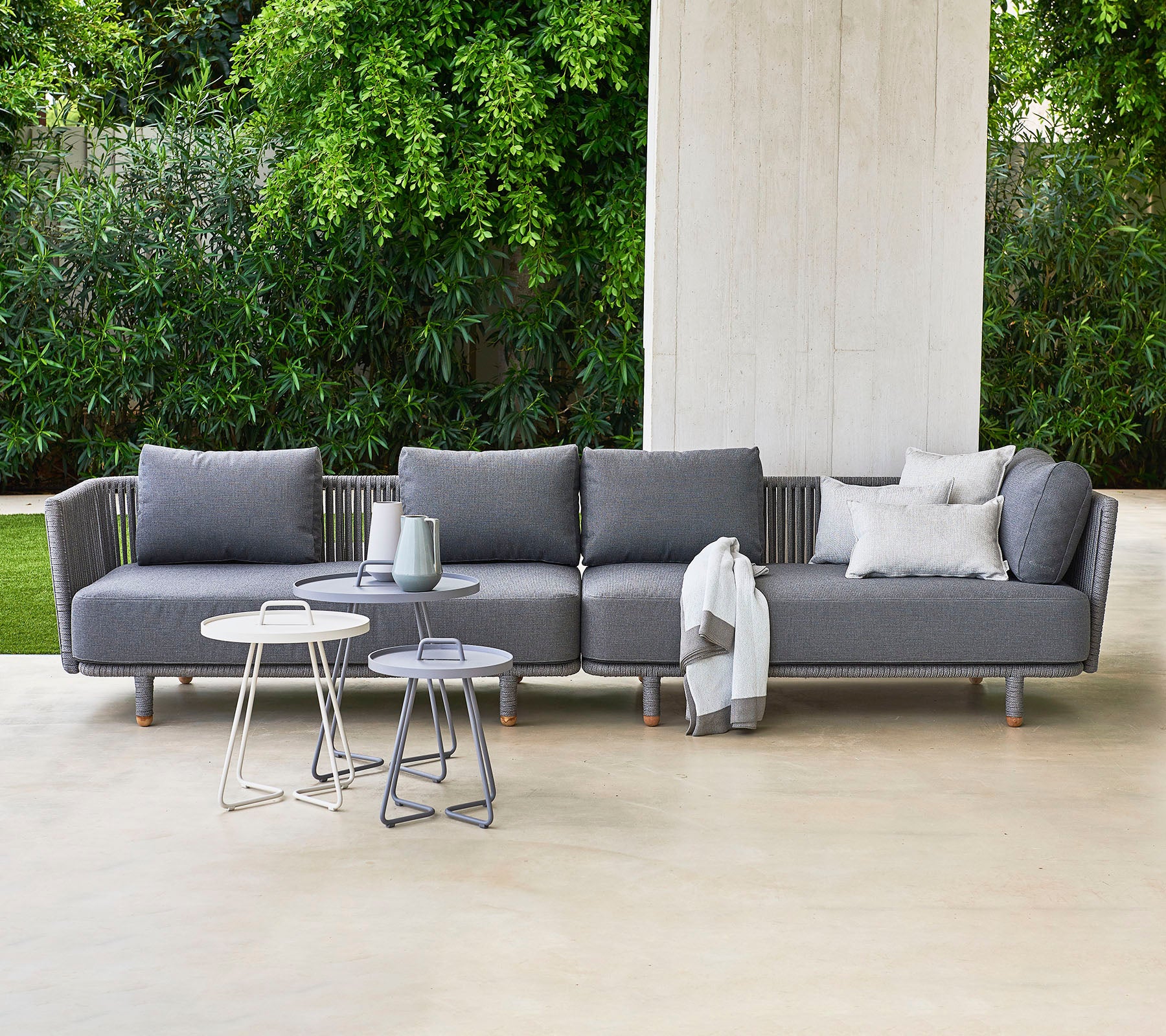 Canapé sectionnel gris moderne avec des coussins, accompagné de deux tables d'appoint élégantes, sur fond de verdure luxuriante.