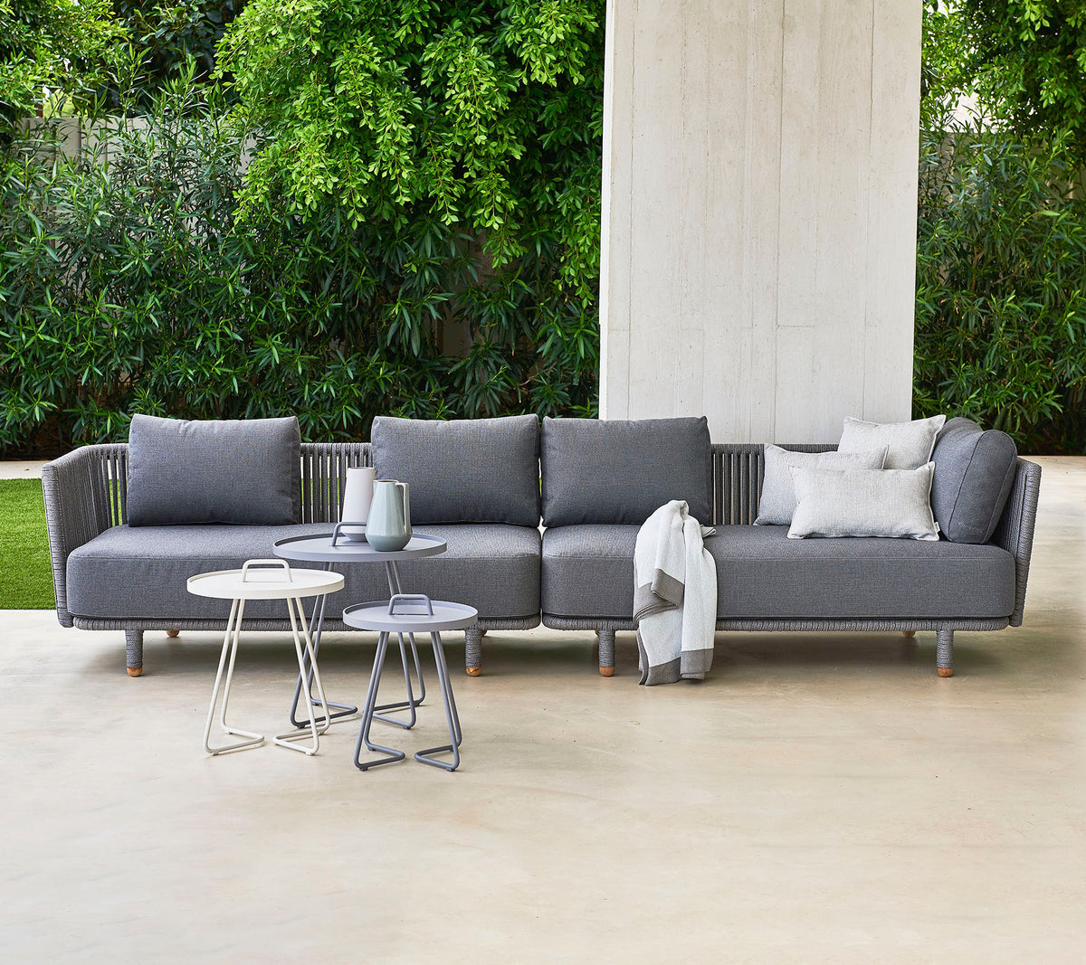 Canapé sectionnel gris moderne avec des coussins, accompagné de deux tables d'appoint élégantes, sur fond de verdure luxuriante.