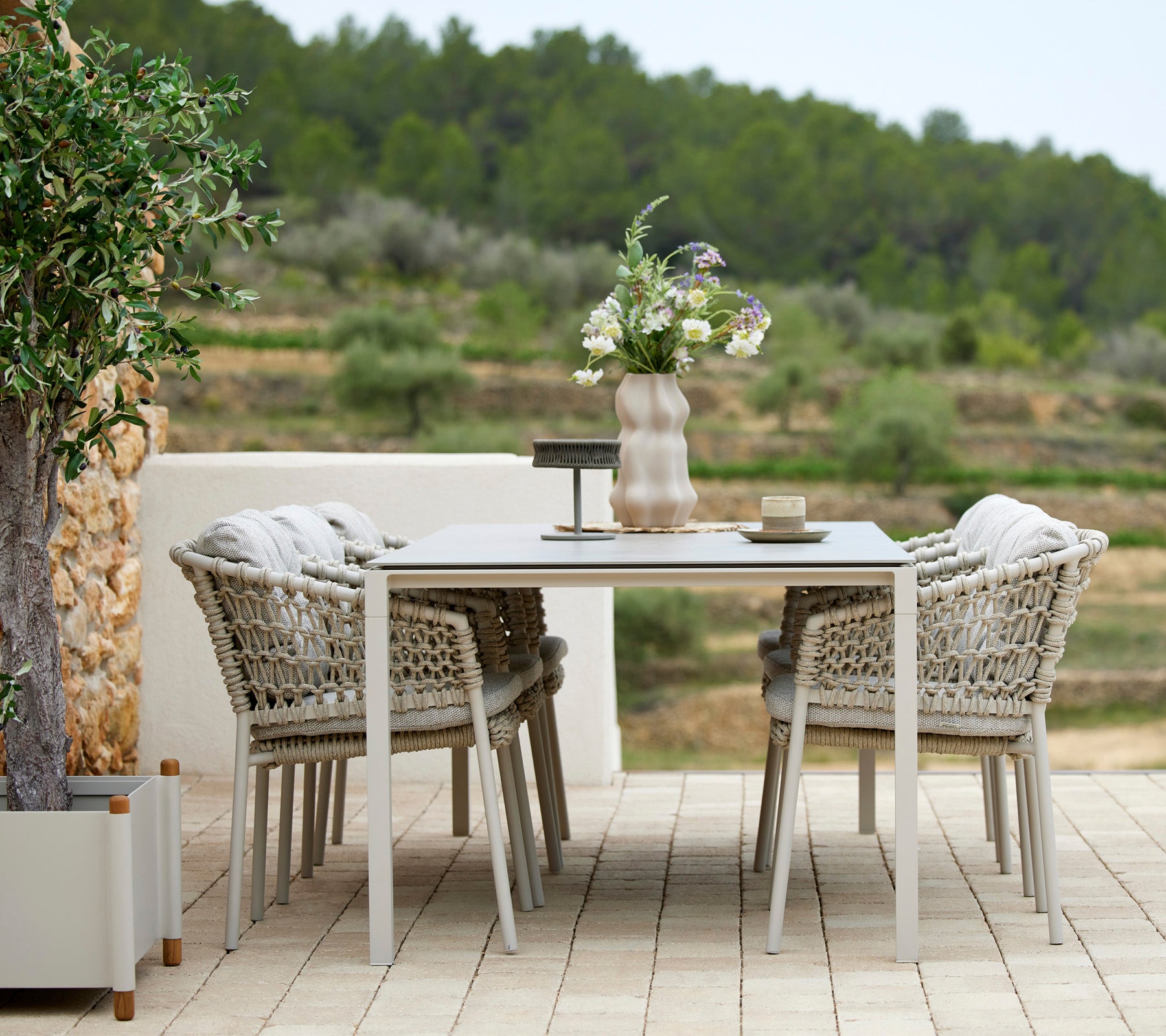 Un ensemble de table à manger d'extérieur élégant avec quatre chaises tressées, décoré d'un vase de fleurs et d'accessoires de table, entouré de verdure.