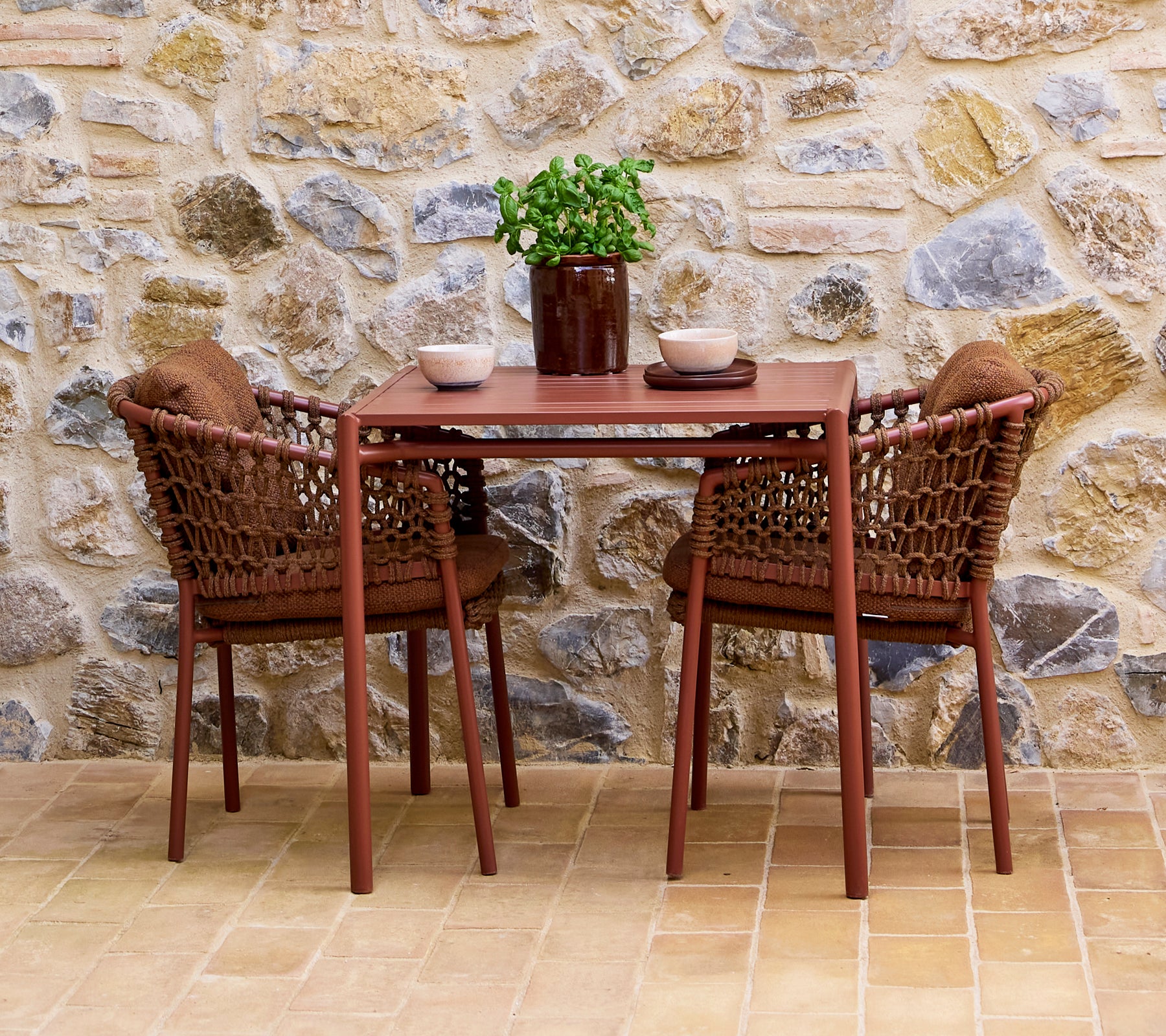 Chaises brun ombre assorties avec une table assortie, accentuées par une plante en pot et des bols décoratifs, devant un mur en pierre.