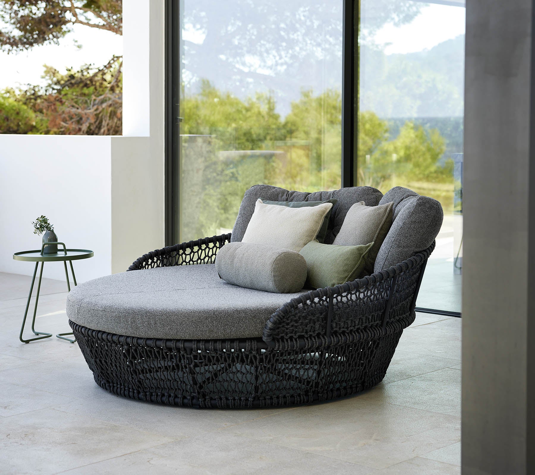 Lit de jour élégant avec des coussins gris, entouré de grandes fenêtres offrant une vue extérieure pittoresque.