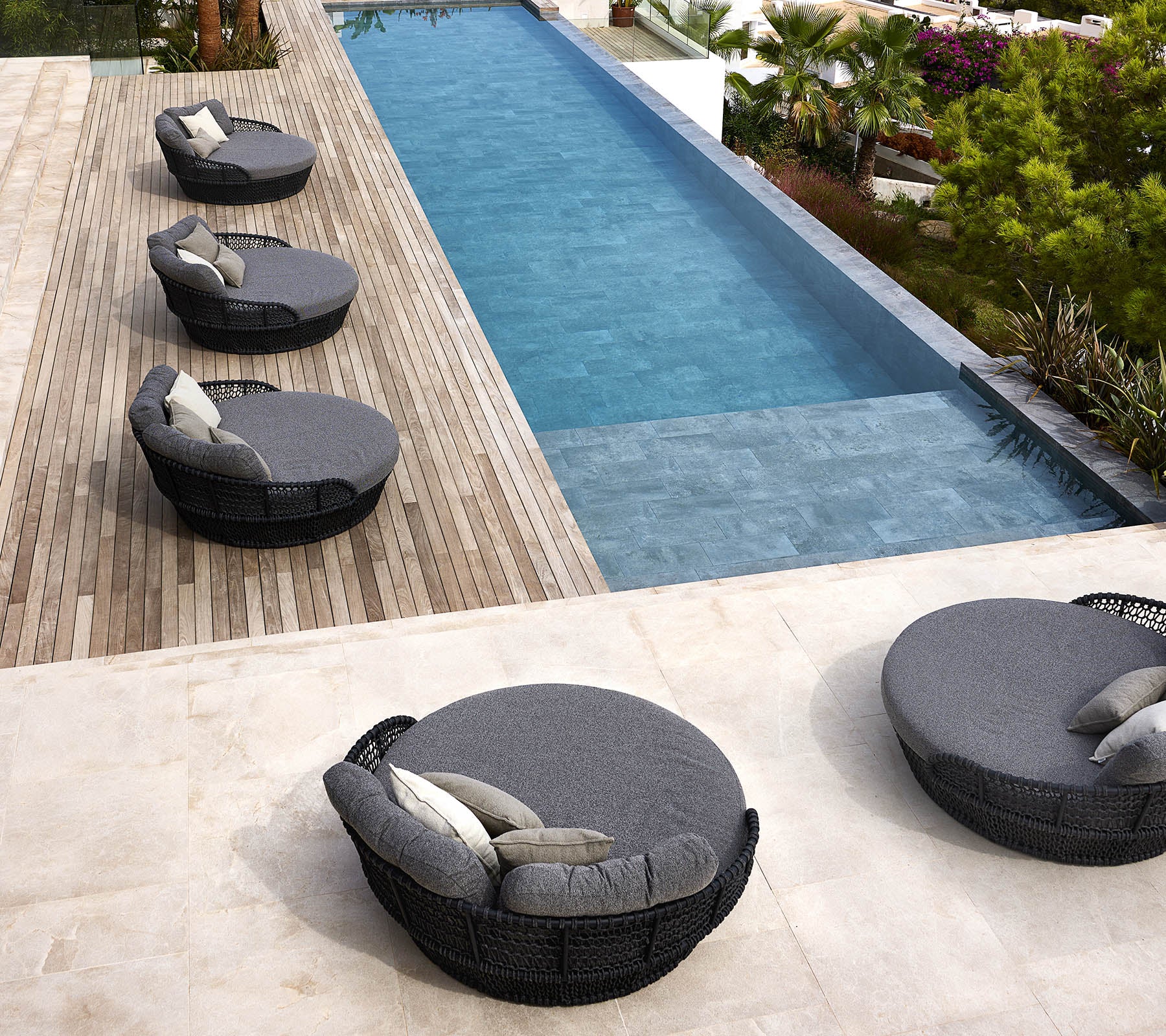 Quatre lits ronds de jour en bord de mer avec des coussins sont disposés à côté d'une piscine élégante entourée de verdure et de terrasses en bois.
